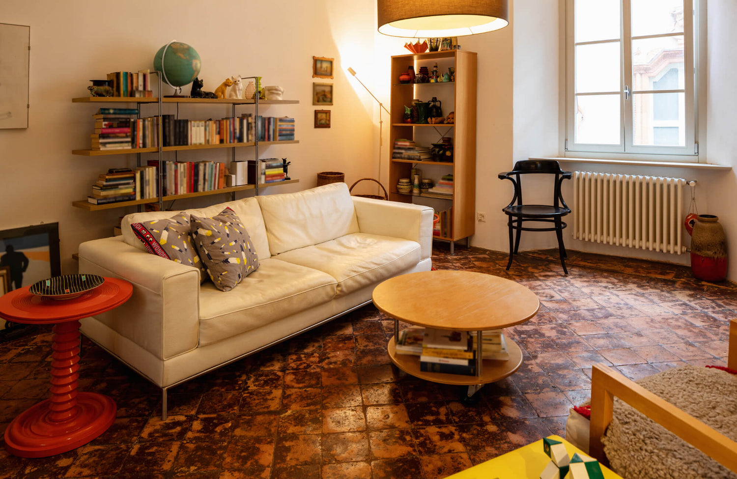 Cozy living room with terracotta tile flooring, white sofa, bookshelves, modern lighting, and eclectic decorative accents.