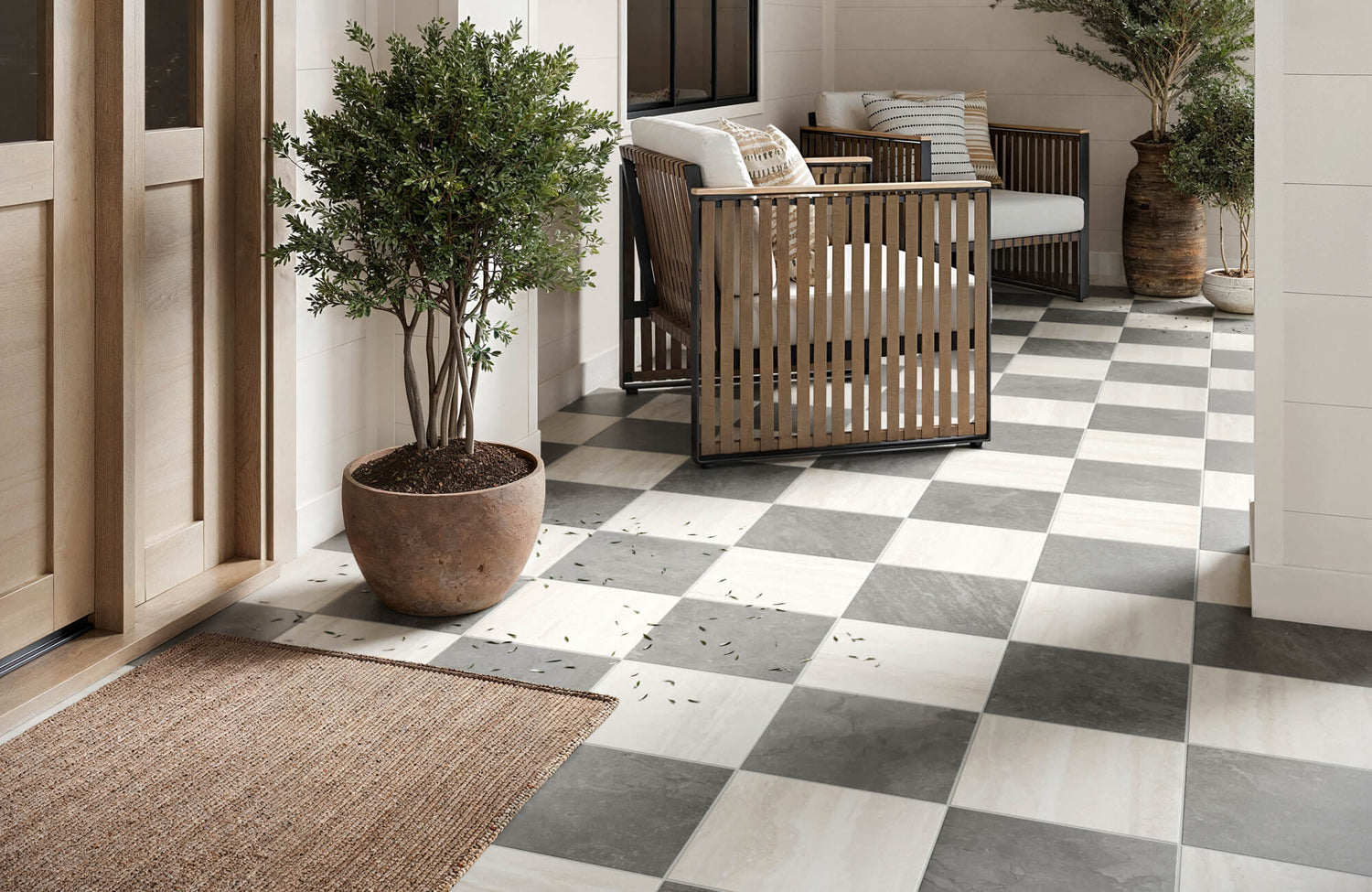 Outdoor patio with classic gray and white checkerboard tile flooring leading to a seating nook with wood lounge chairs, soft cushions, and potted plants.