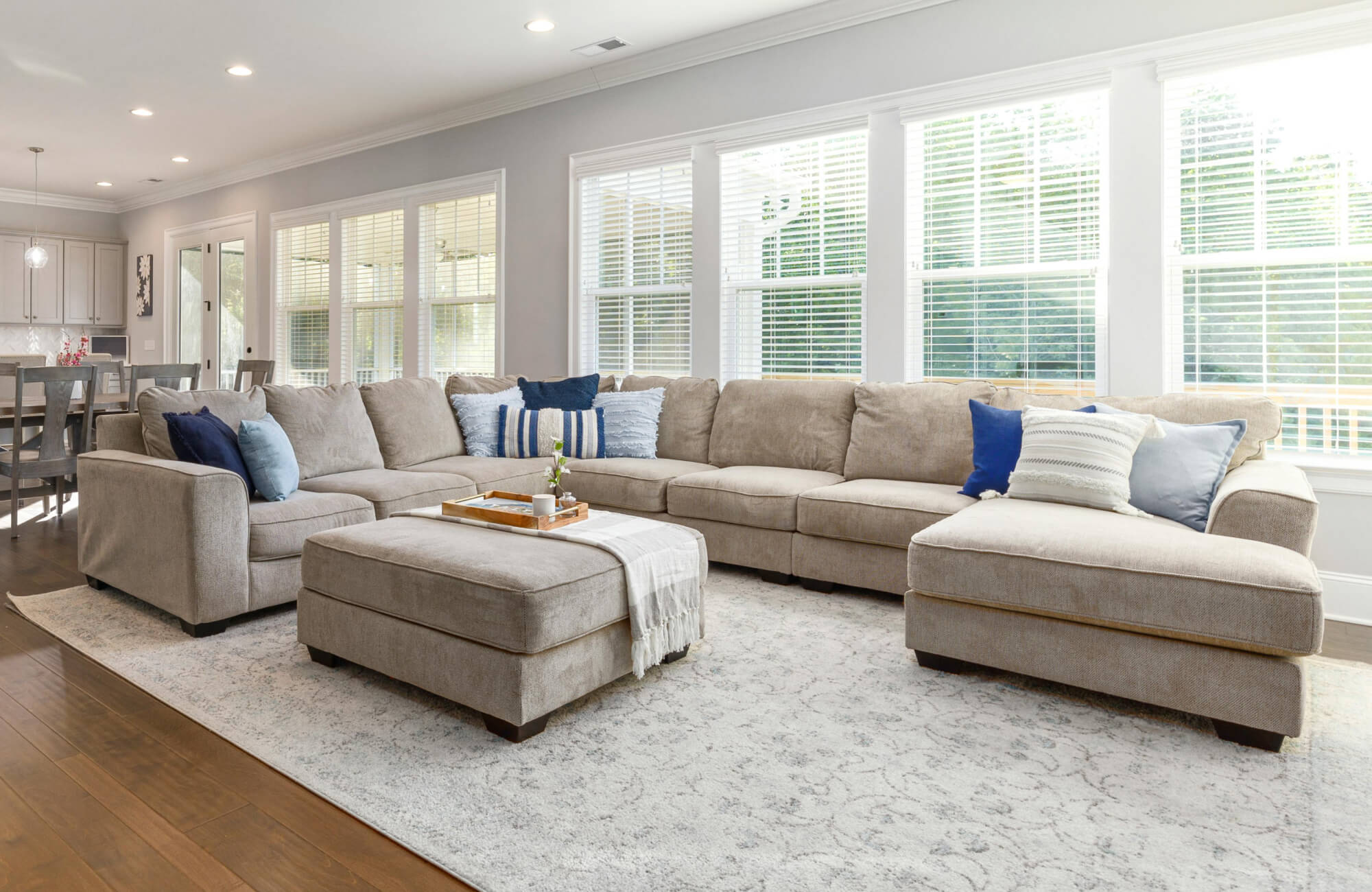 Contemporary open-concept living room with a large beige sectional sofa, matching ottoman, blue and white throw pillows, light patterned area rug, wood flooring, and expansive windows bringing in natural light.