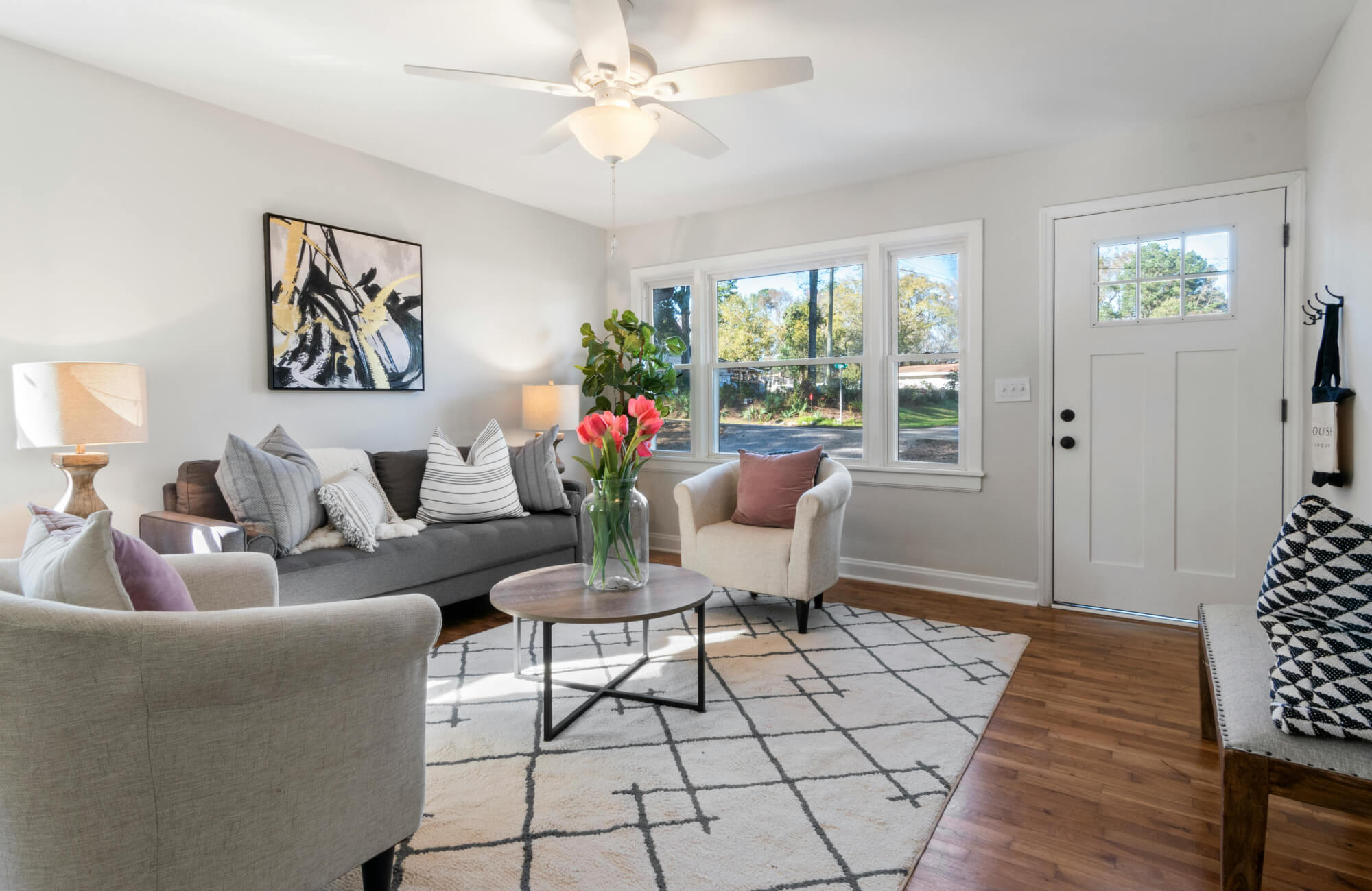 Bright contemporary living room with patterned geometric rug, neutral seating with decorative pillows, wall art, desk lamp, pendant lights, wood flooring, and airy entryway design with natural light. 