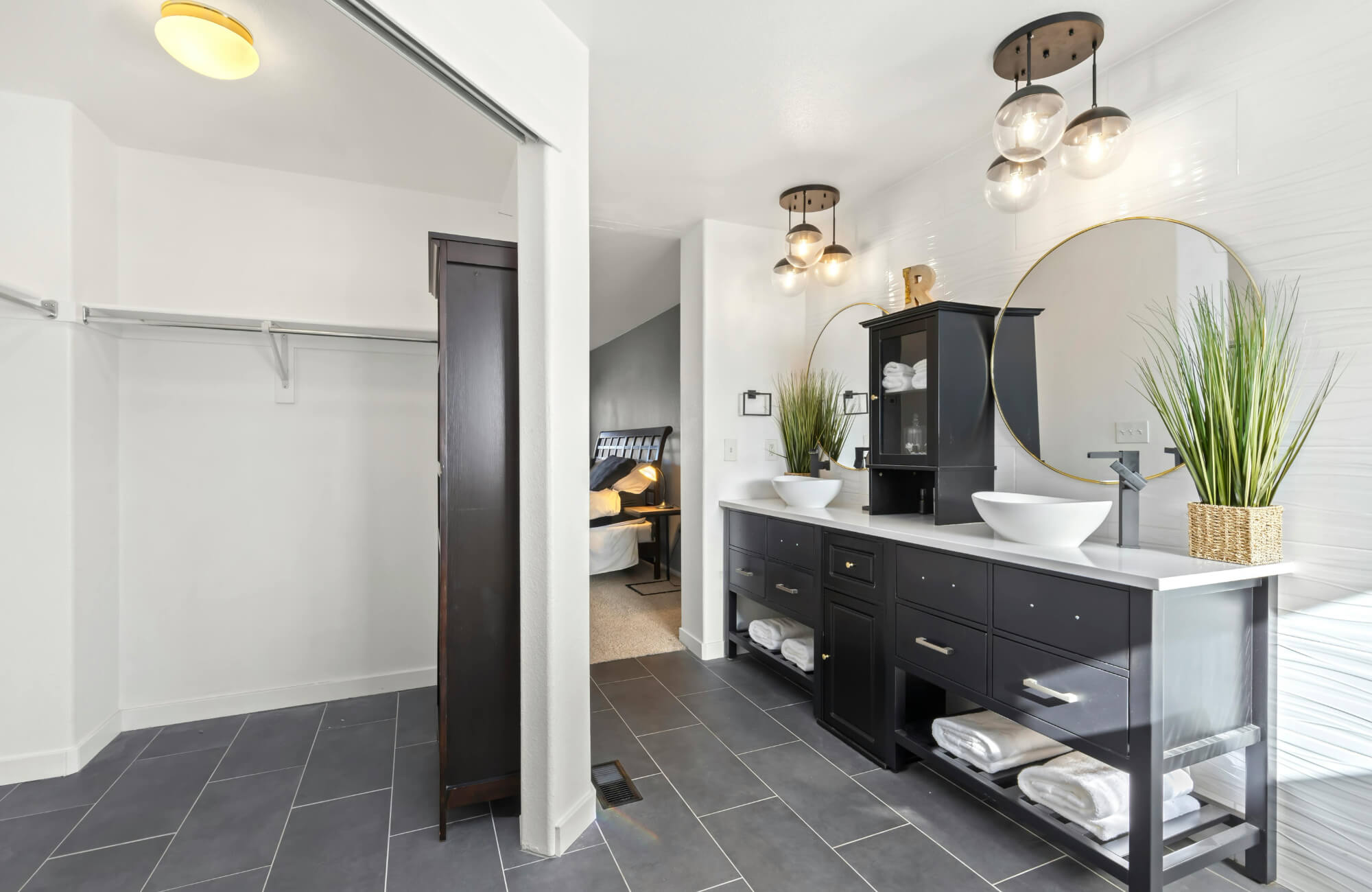 Modern bathroom with a black double sink vanity, vessel sinks, round mirrors, and open shelving, adjacent to a walk-in closet.