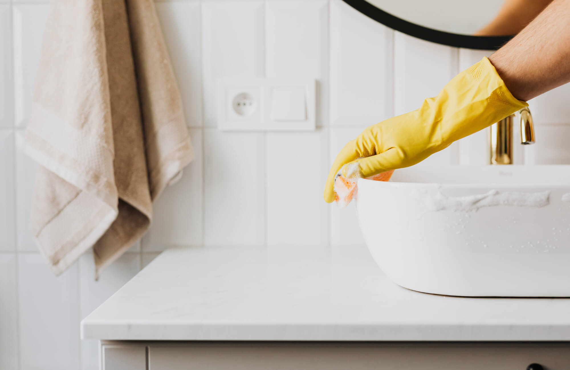 A person wearing a yellow cleaning glove wiping a white bathroom sink with a sponge on a quartz countertop in a modern, tiled vanity setup.