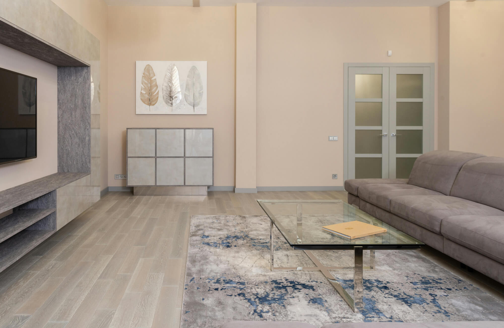 Modern living room with a gray abstract rug featuring blue accents, a glass coffee table, a beige sofa, light wood floors, and minimalist decor on soft beige walls.