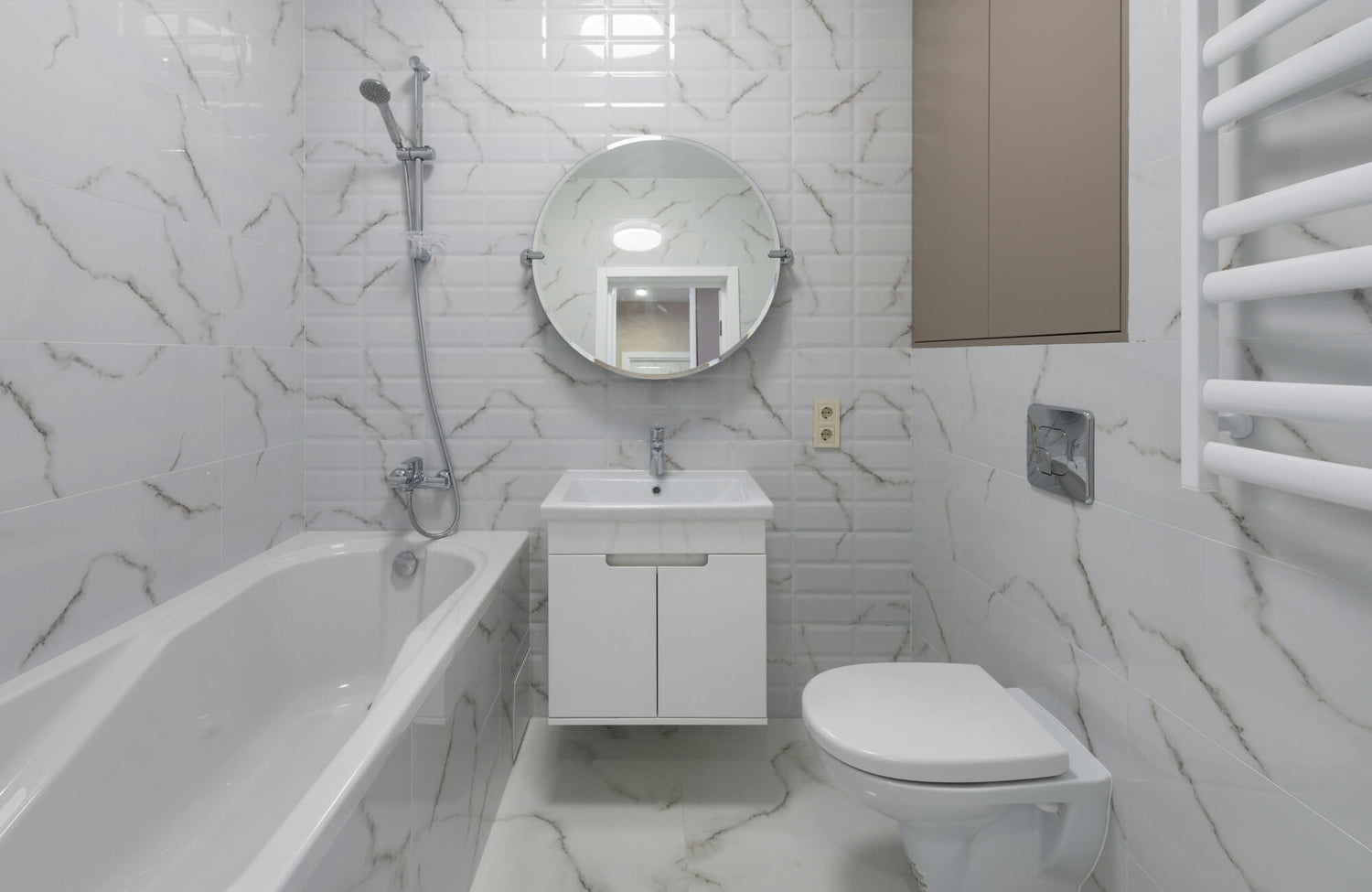 Compact bathroom with white marble-effect tiles, wall-mounted vanity, round mirror, and built-in tub with handheld shower.