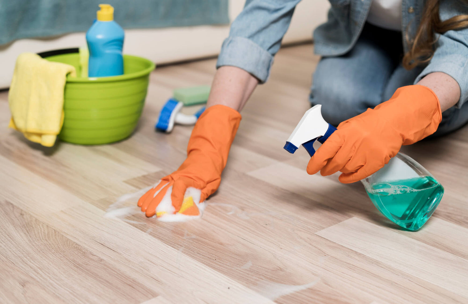 A person wearing orange gloves is scrubbing vinyl plank flooring with a sponge and cleaning spray, with a green bucket and cleaning supplies nearby.