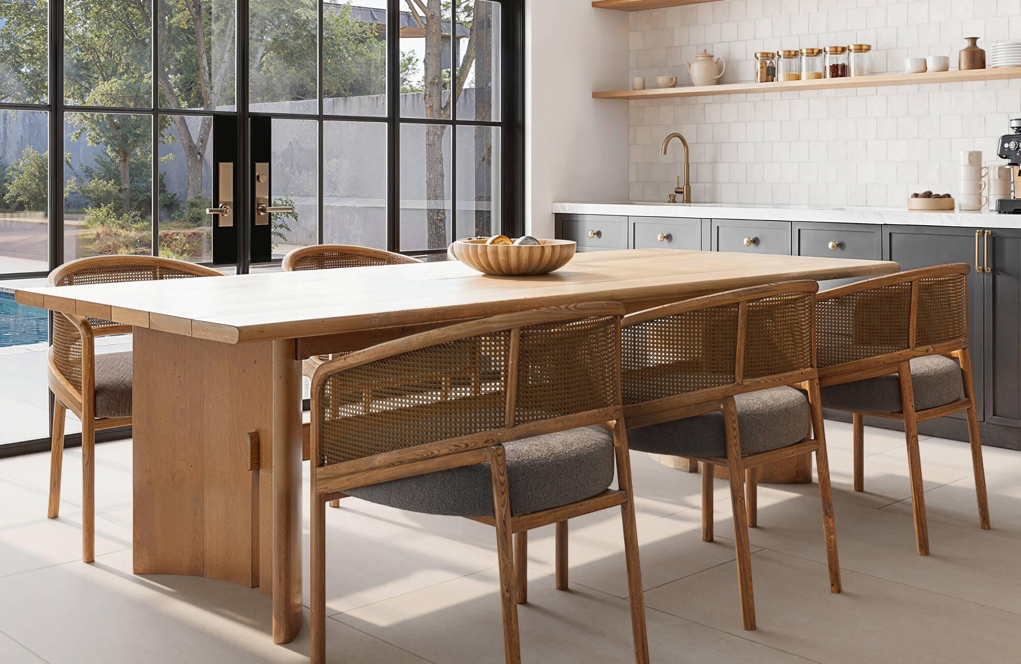 Modern dining table with a light wood finish and clean rectangular design, complemented by woven-back chairs and floor-to-ceiling windows that fill the airy kitchen with natural light and warmth.