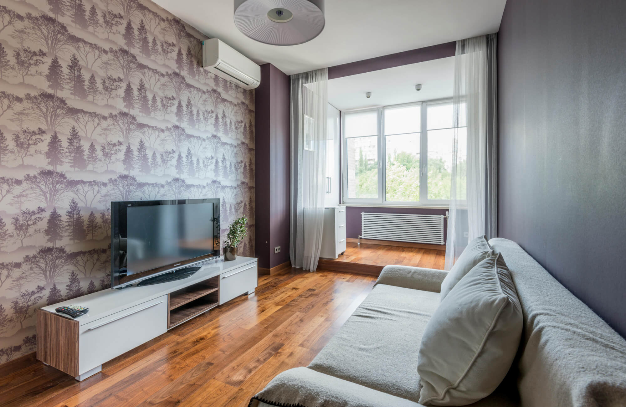 Modern living room featuring forest-patterned wallpaper in muted mauve tones, paired with a gray sofa, wooden flooring, and a minimalist TV console.