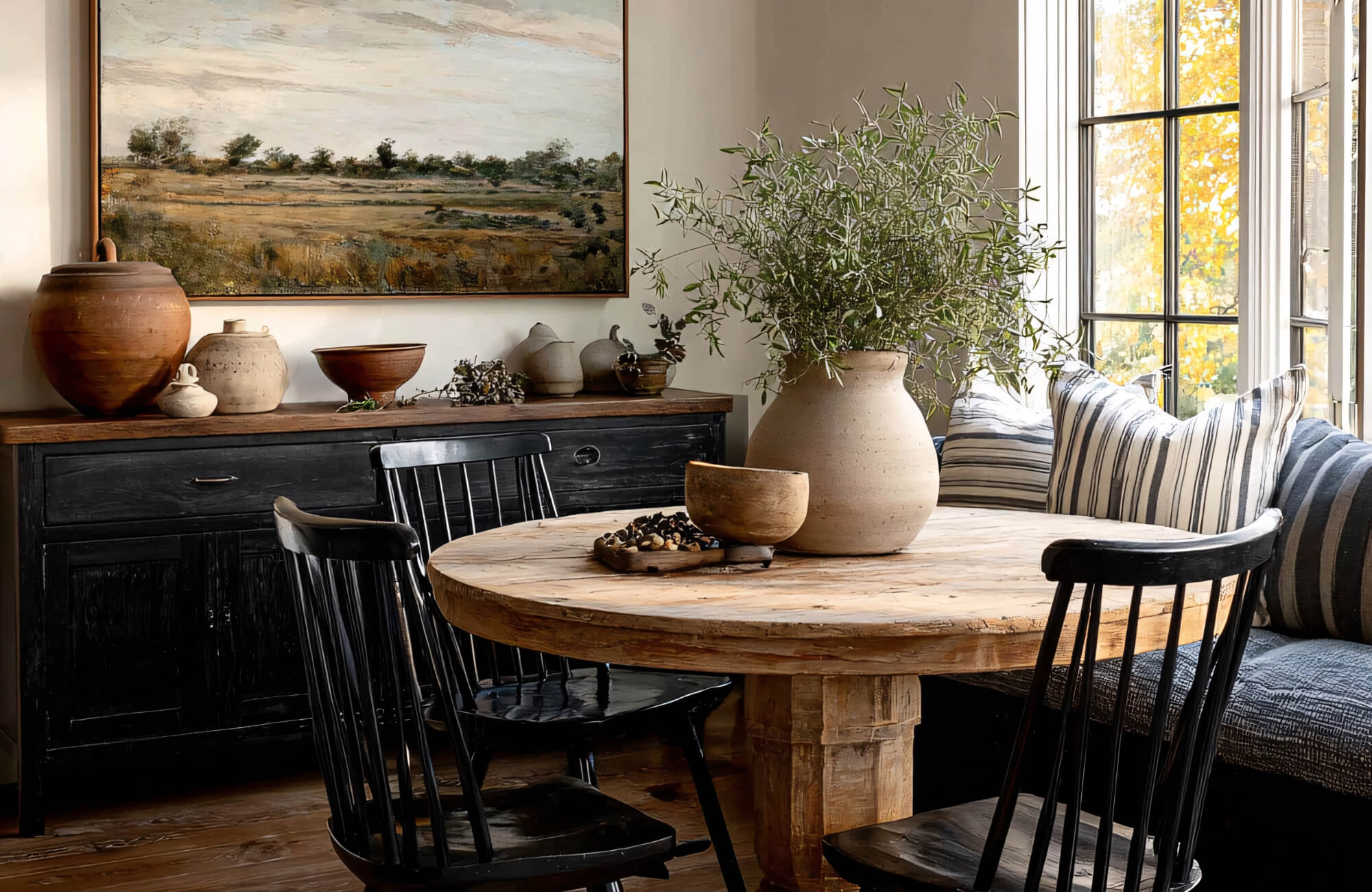 Black dining chairs paired with a light wood round table, styled with pottery and greenery to create warmth and contrast in a bright dining nook.