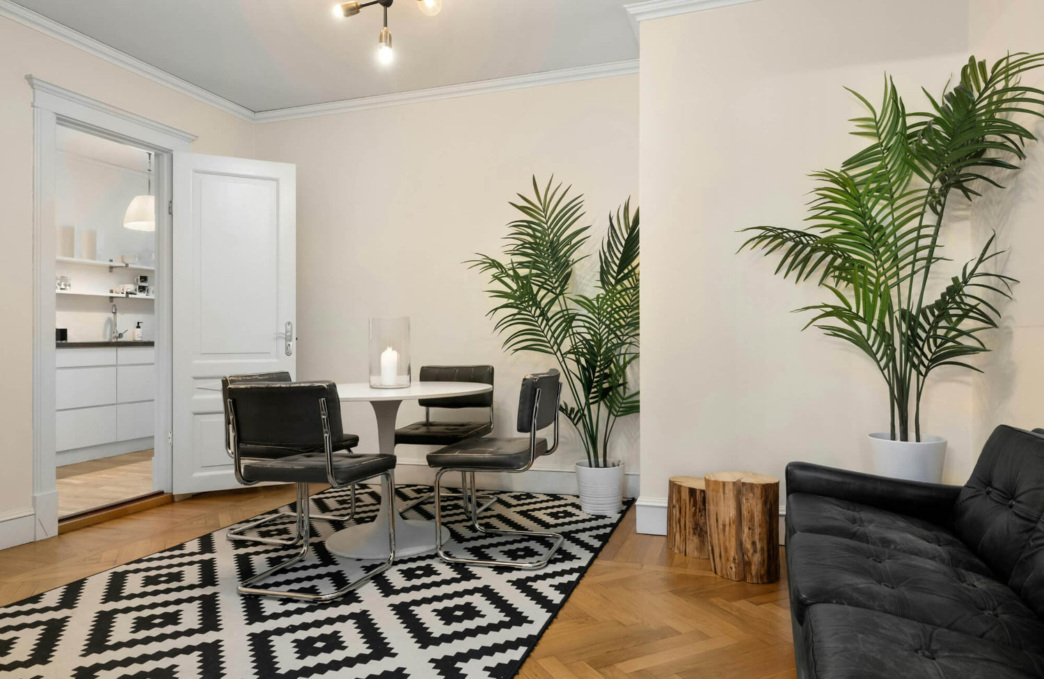 Modern dining area with black-and-white geometric rug, round table, black chairs, potted palms, and black leather sofa.