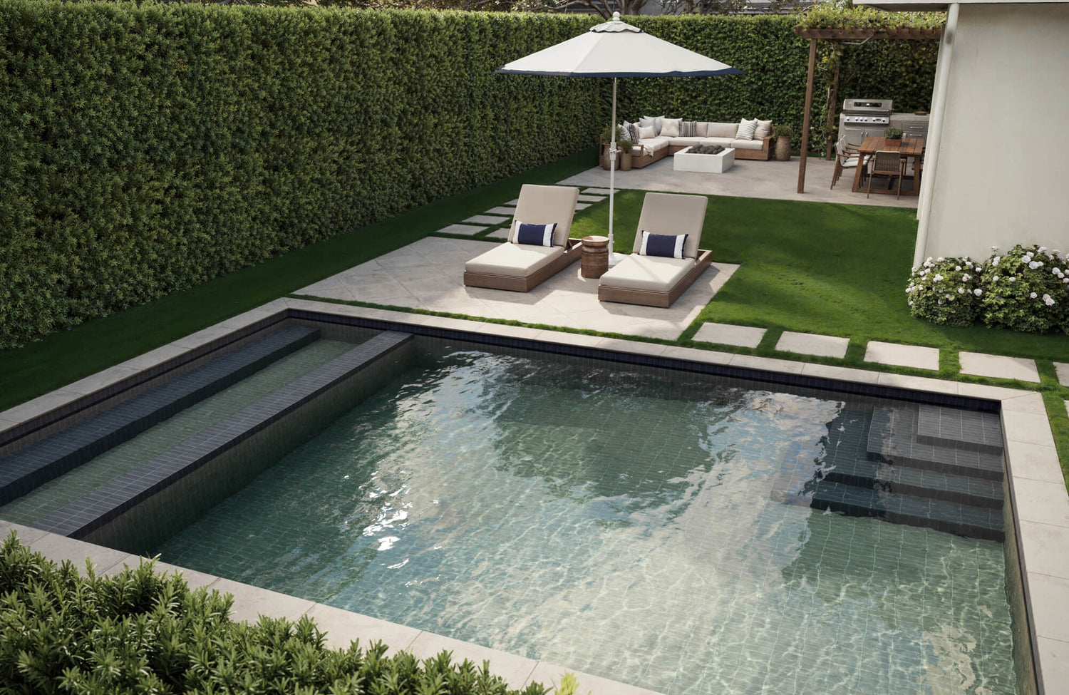 Modern backyard with a white paver tile poolside, cushioned loungers, and an outdoor lounge area with grill and fire pit.