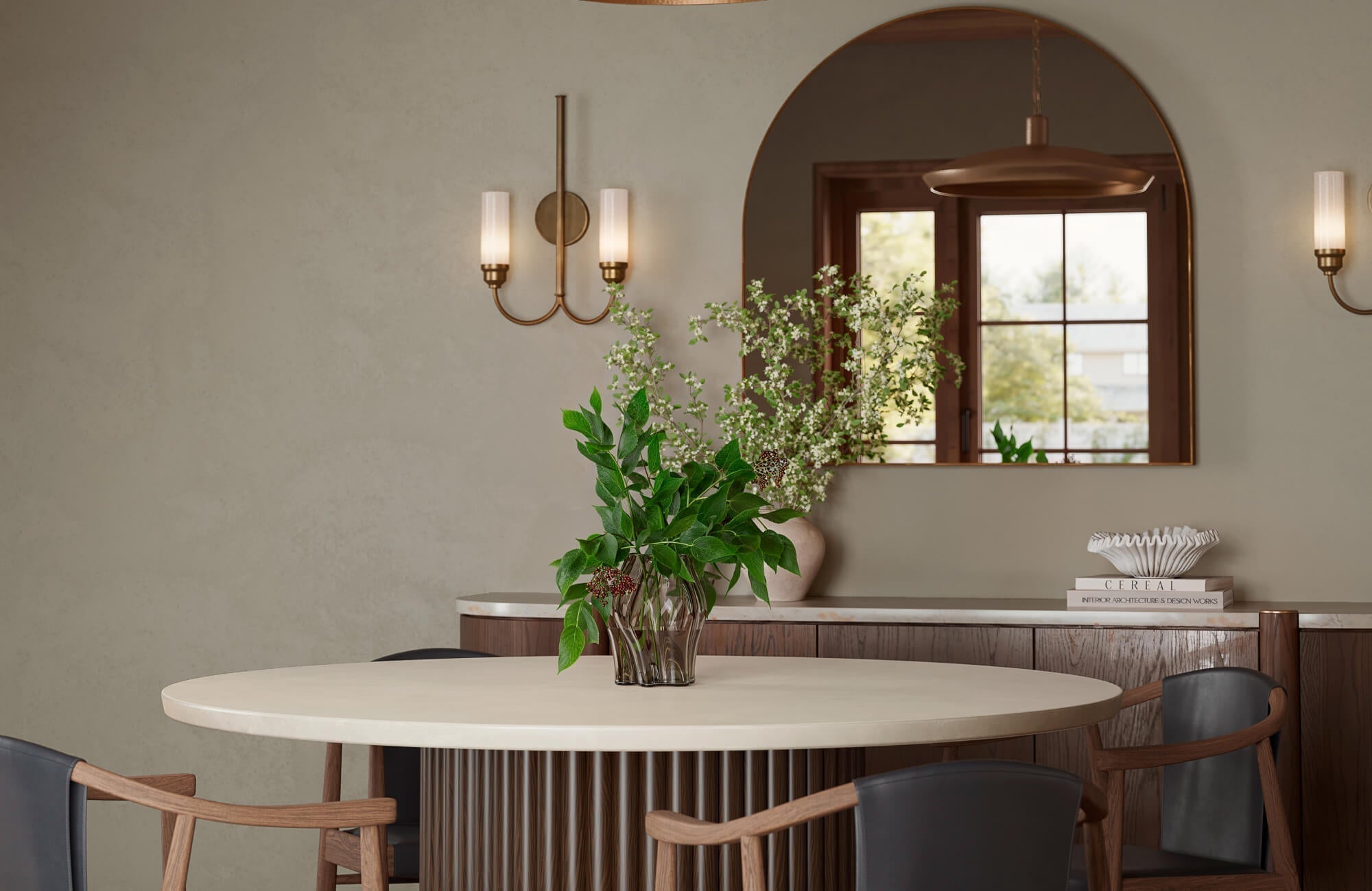 Elegant dining nook with a round fluted-base table, curved wood chairs, an arched wall mirror, brass sconces, and fresh greenery.