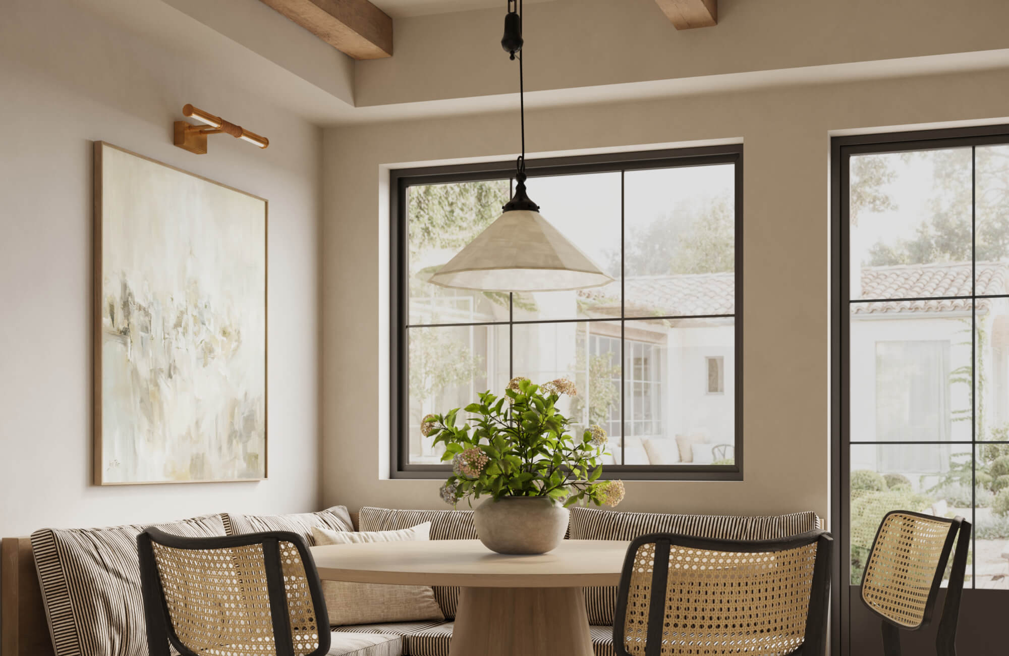 Cozy dining nook with picture lights above abstract art, a round wooden table, woven chairs, and a potted plant by large windows.