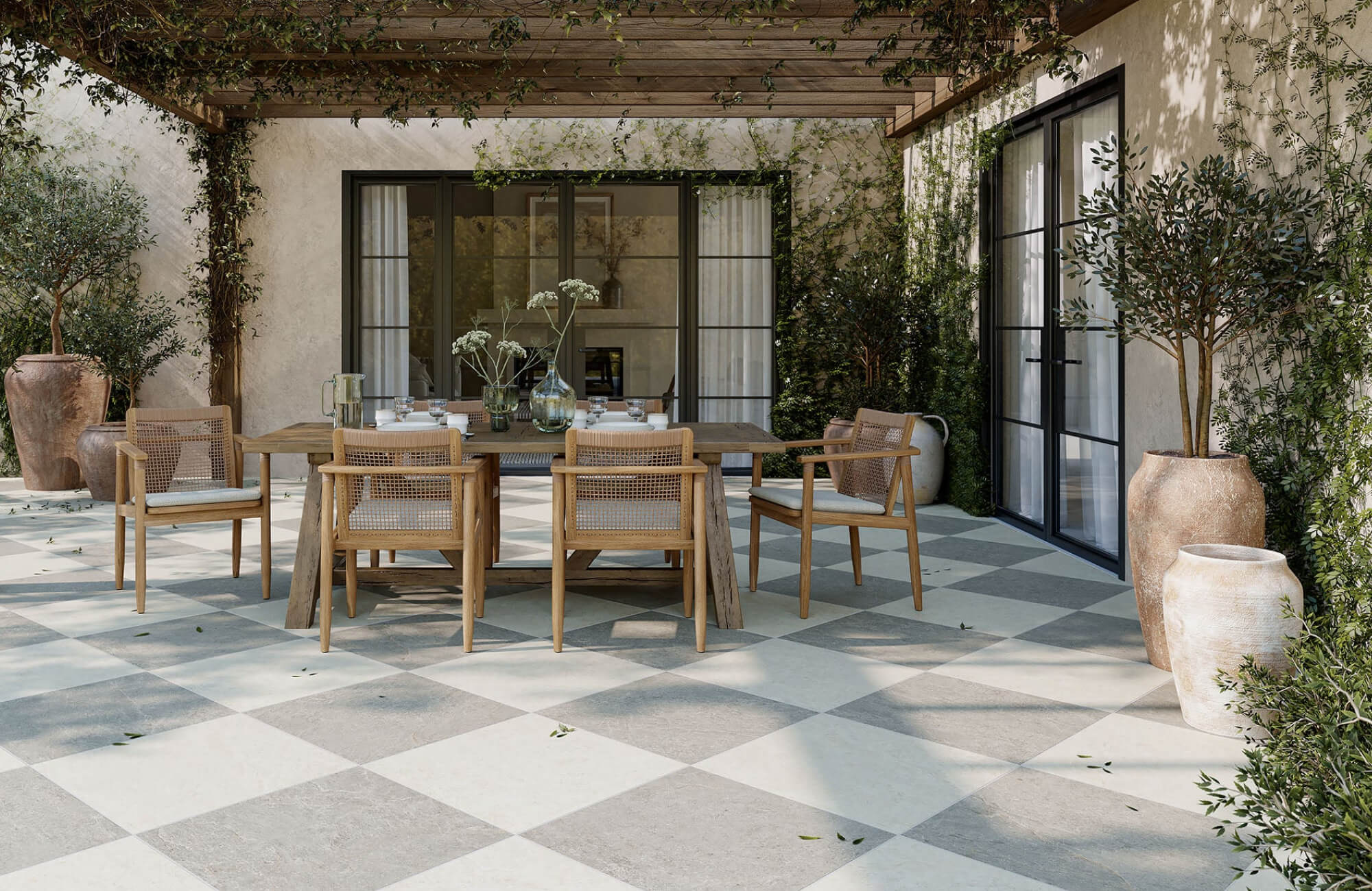 Outdoor patio dining area with a rectangular wooden table, woven armchairs, large planters, glass doors, and a shaded pergola covered in greenery.