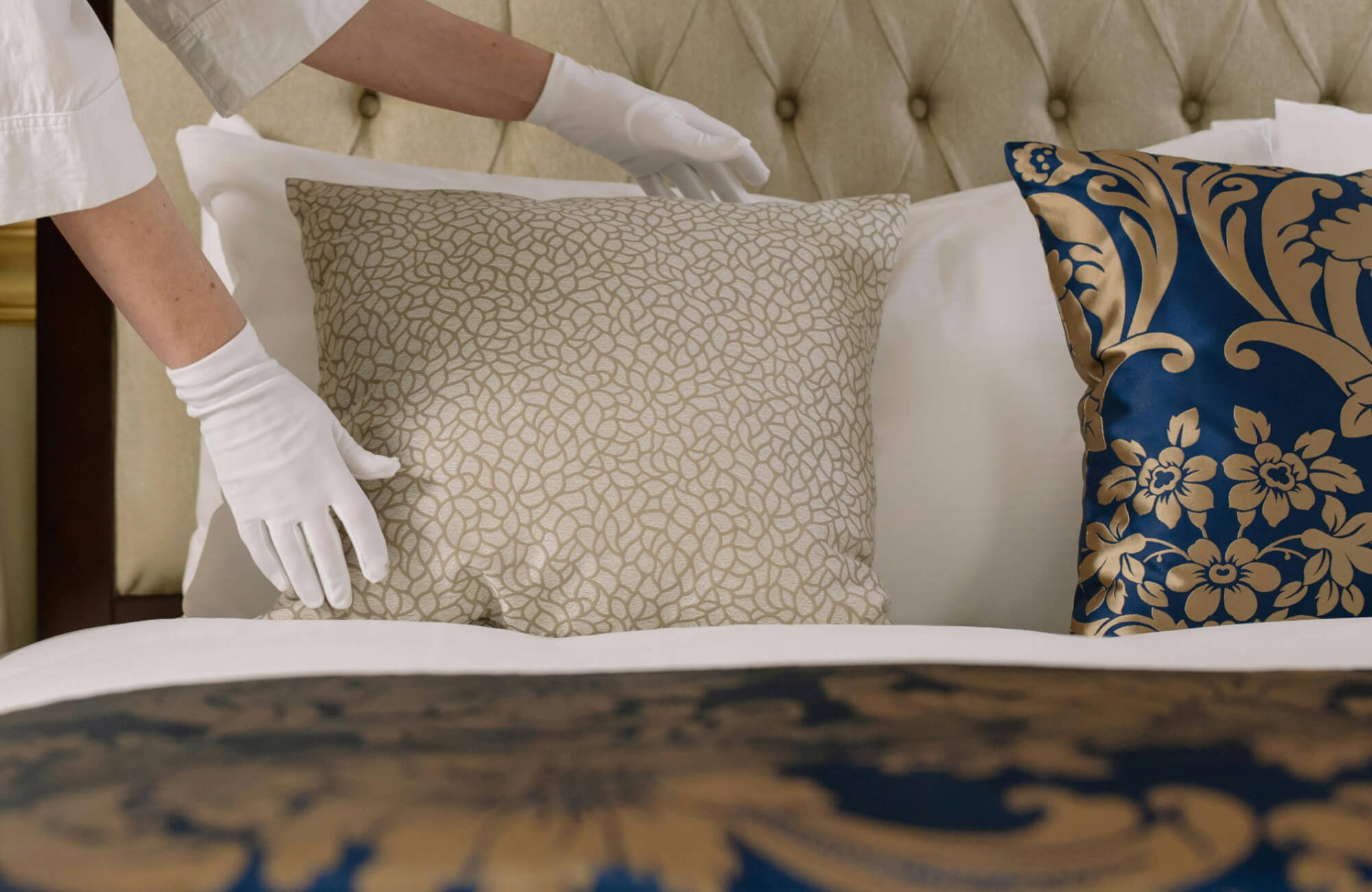 Gloved hands arranging patterned decorative pillows on a plush bed with a tufted headboard and elegant blue and gold accents.