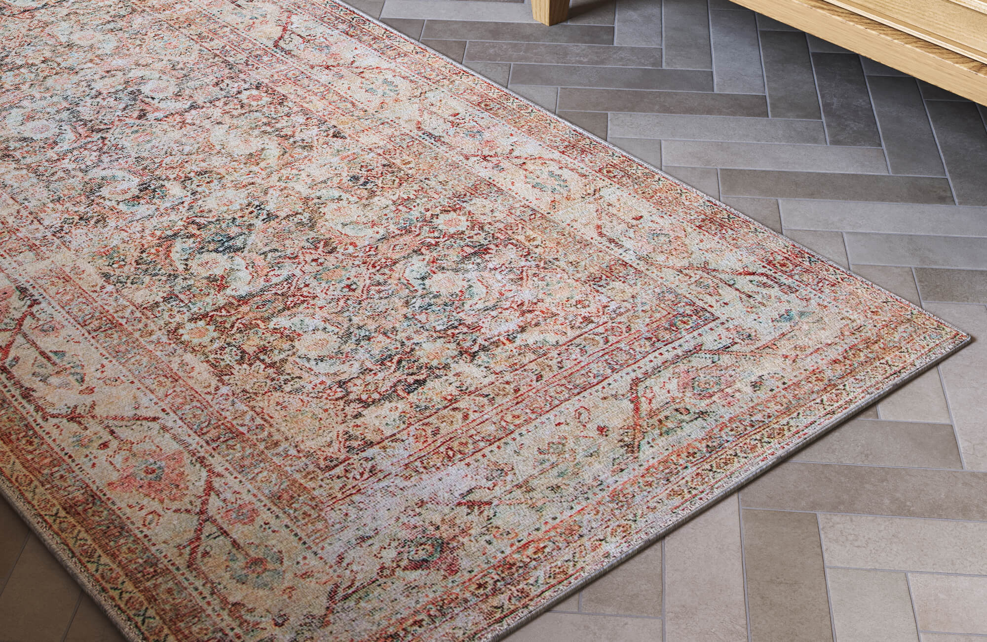 Close-up of a faded traditional area rug with red, beige, blue, and gray detailing laid over gray herringbone tile flooring beside a wood vanity.