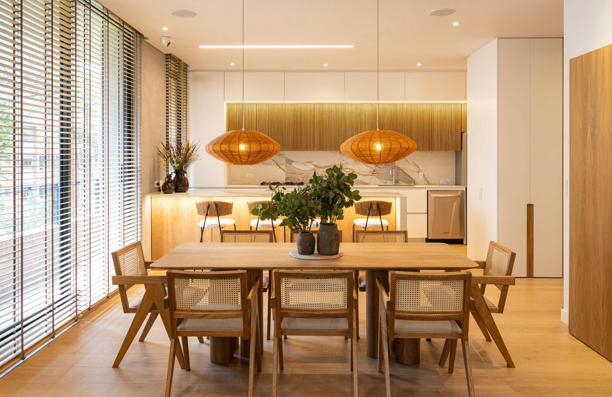 Warm woven pendant lights illuminate a natural wood dining table in a bright, modern dining room.