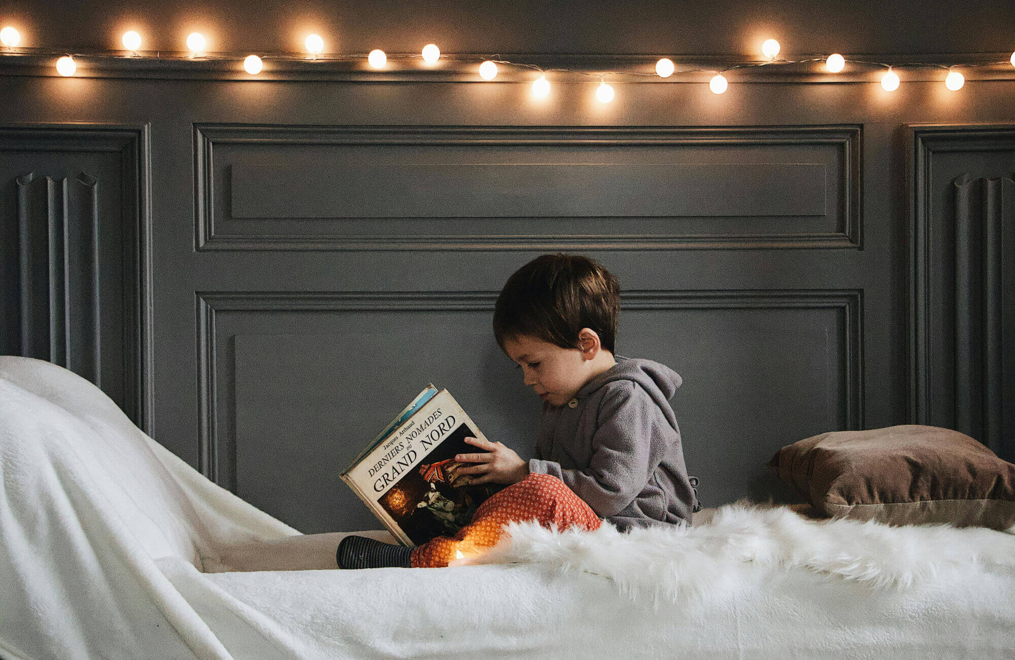 A kid sitting on a bed with warm fairy lights above, enjoying a quiet moment in a softly lit room.