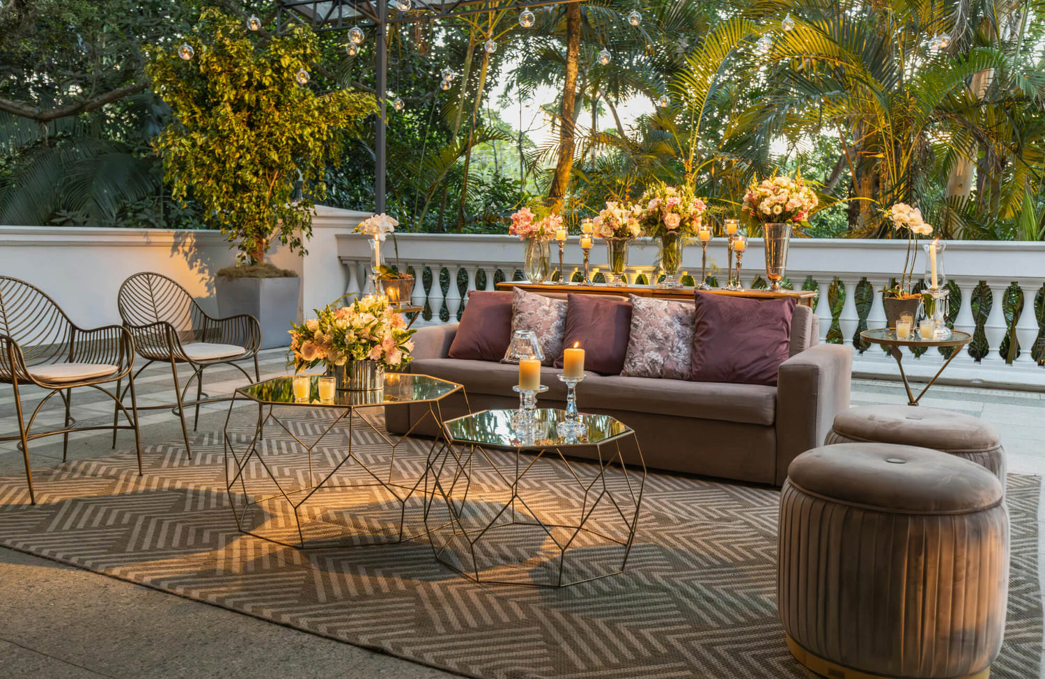 Outdoor lounge with taupe sofa, plum pillows, gold glass tables, floral decor, and candles on a beige herringbone rug under string lights and lush greenery.