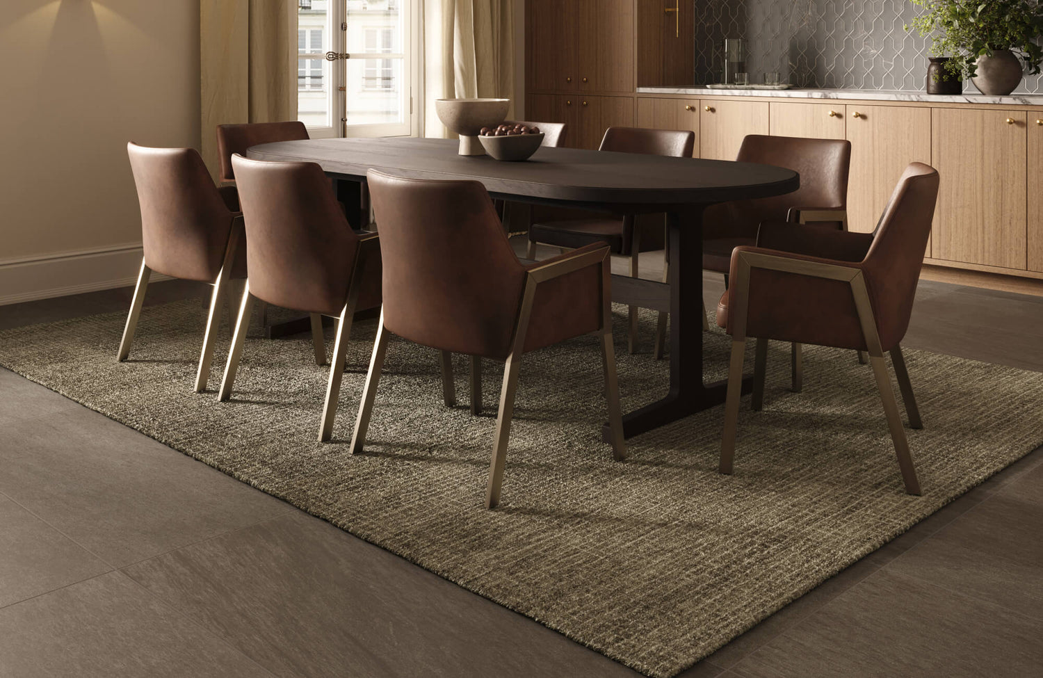 A large brown and beige textured wool rug anchors a dark wood oval dining table with eight brown leather chairs on a dark gray tile floor.