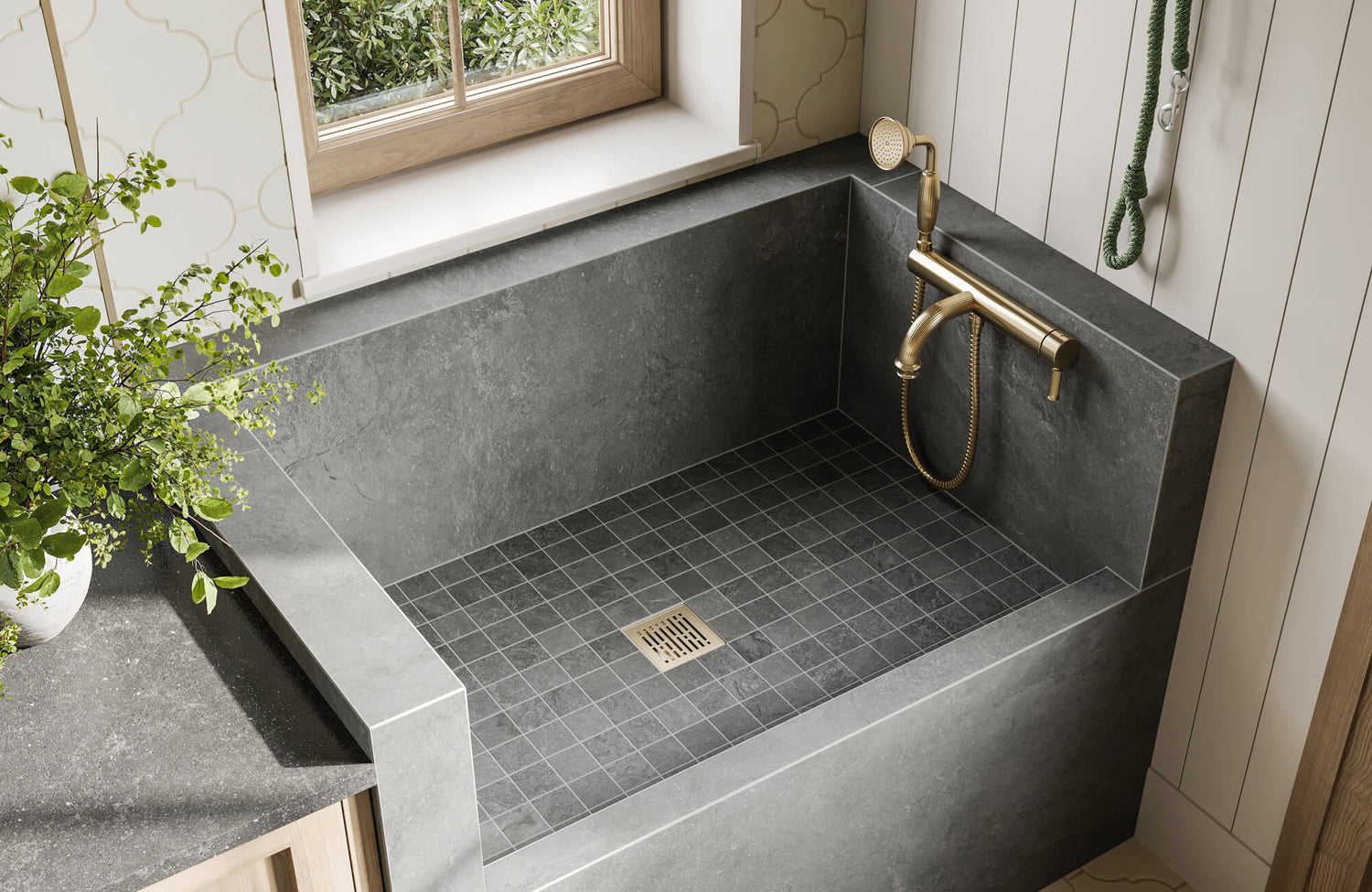 Gray stone tub lined with small square mosaic tiles and accented by a brass handheld shower fixture.