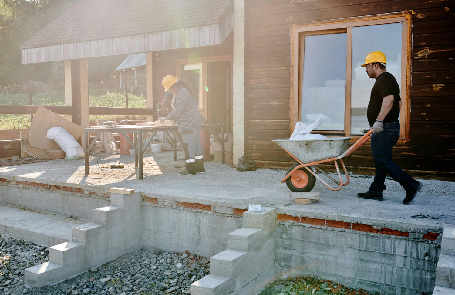 Two workers are building a patio, one transporting supplies in a wheelbarrow while the other measures and cuts materials near a wooden house.