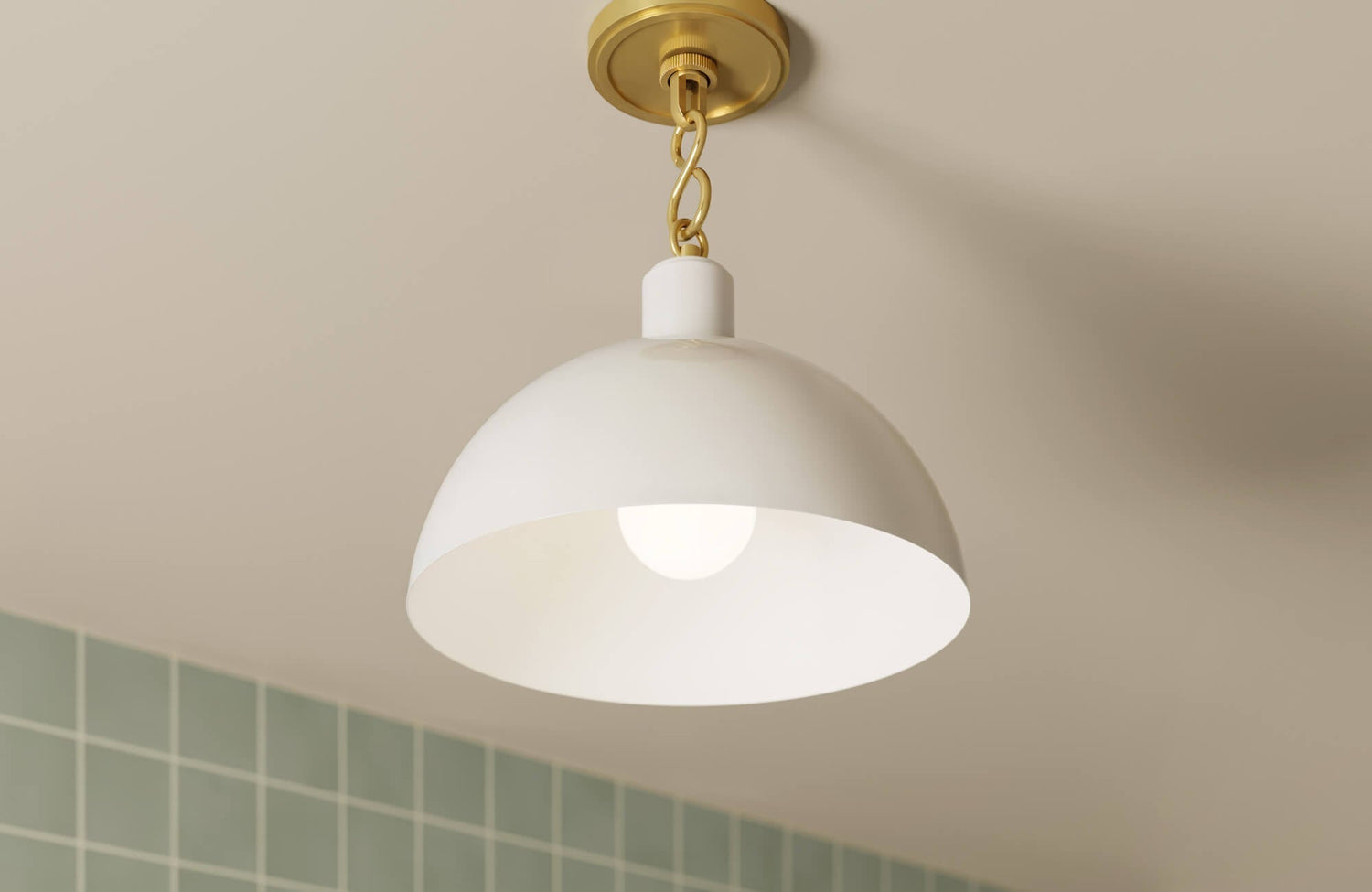 White dome pendant light with a brass chain mount, softly illuminating a space with muted green wall tiles in the background.