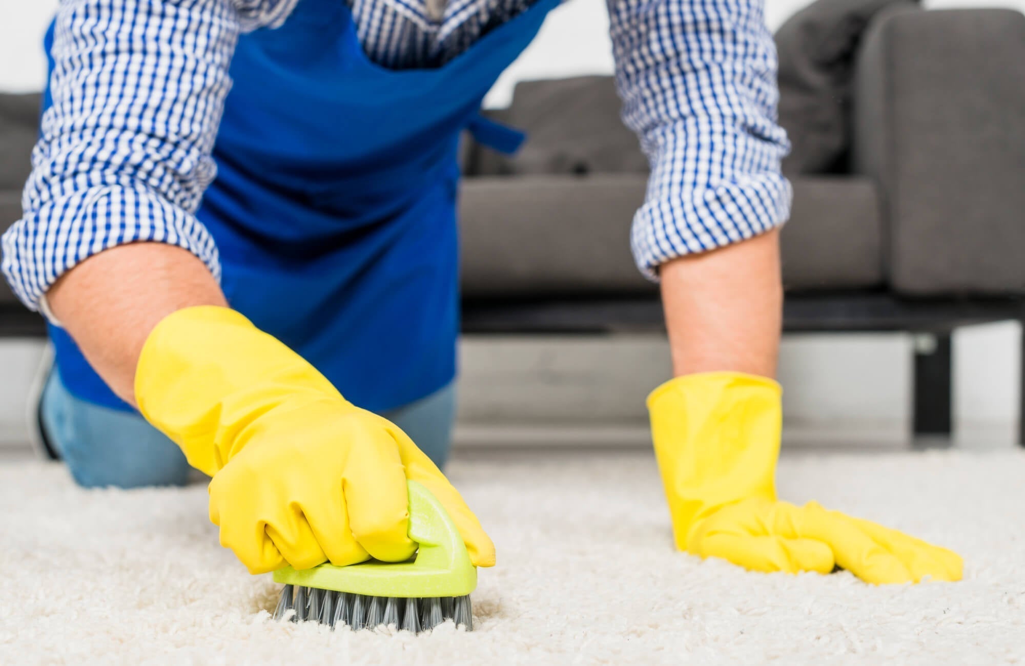 A person wearing yellow gloves is scrubbing a light-colored rug with a brush to remove dirt and stains.