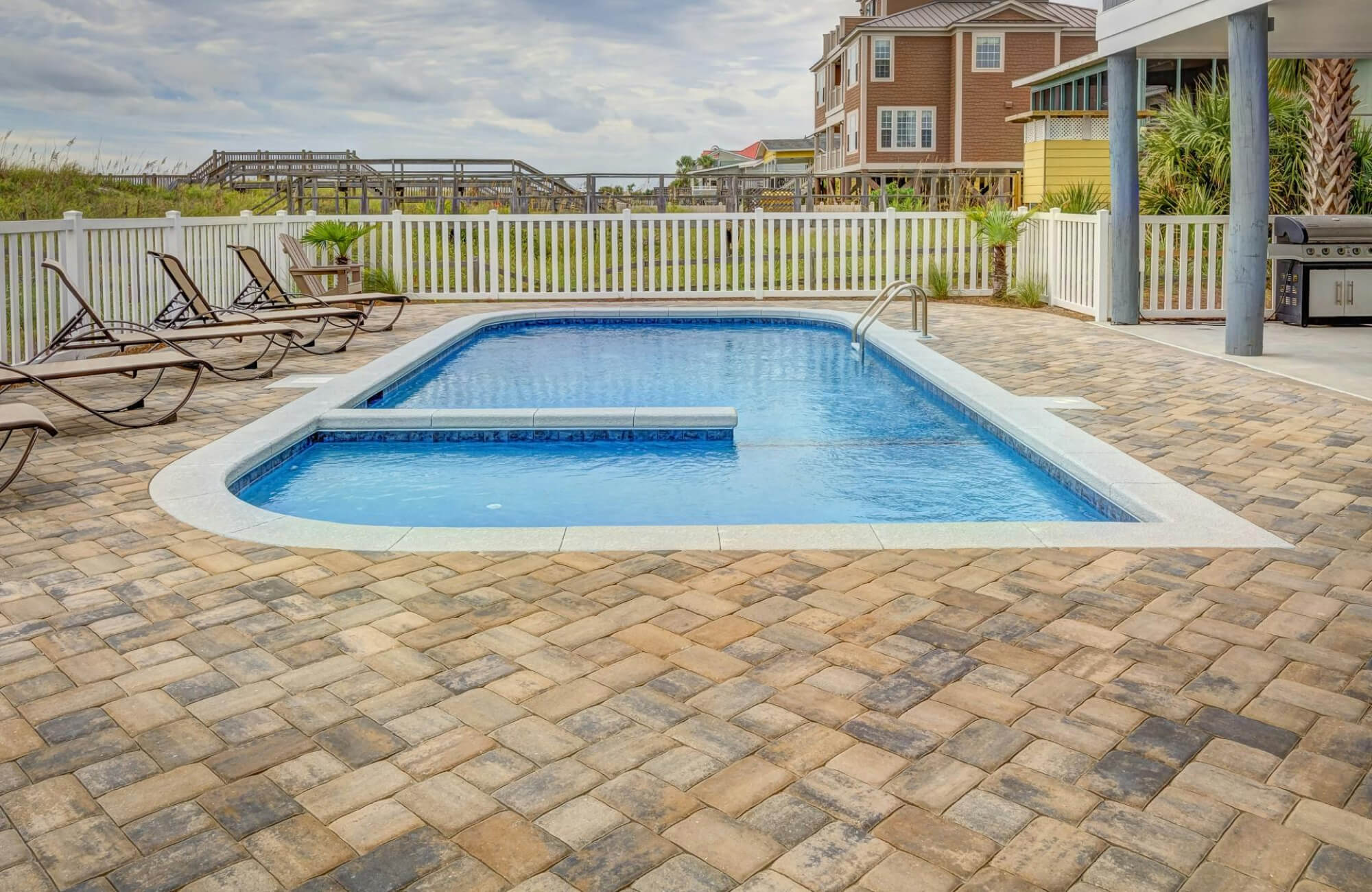 Backyard swimming pool set on multitone paver flooring, featuring several lounge chairs and a white fence overlooking a coastal neighborhood.