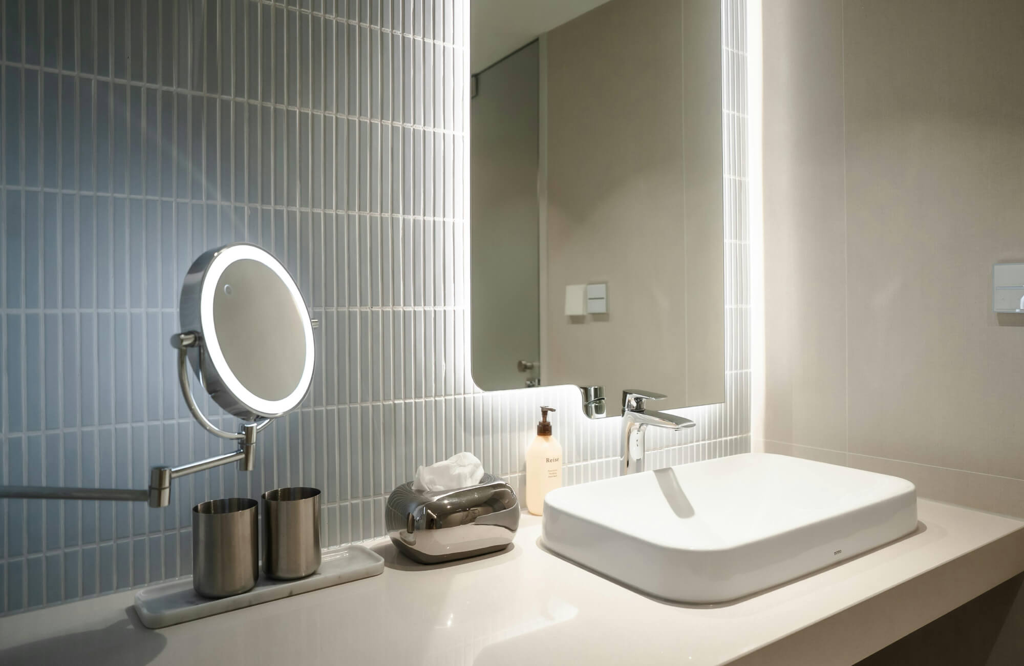 Modern bathroom vanity with a backlit mirror and LED magnifying mirror, paired with vertical blue tile, a sleek basin sink, and soft lighting for daily grooming.