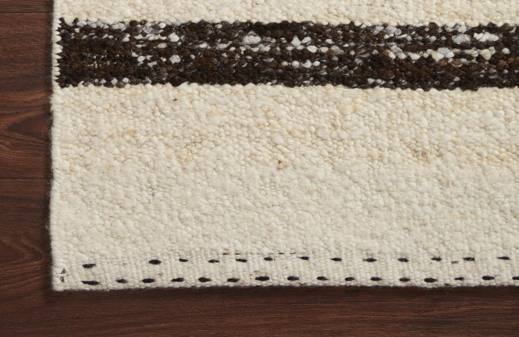 Close-up of a cream textured wool rug with a dark horizontal stripe, stitched border detail, and rich wood flooring visible along the edge.