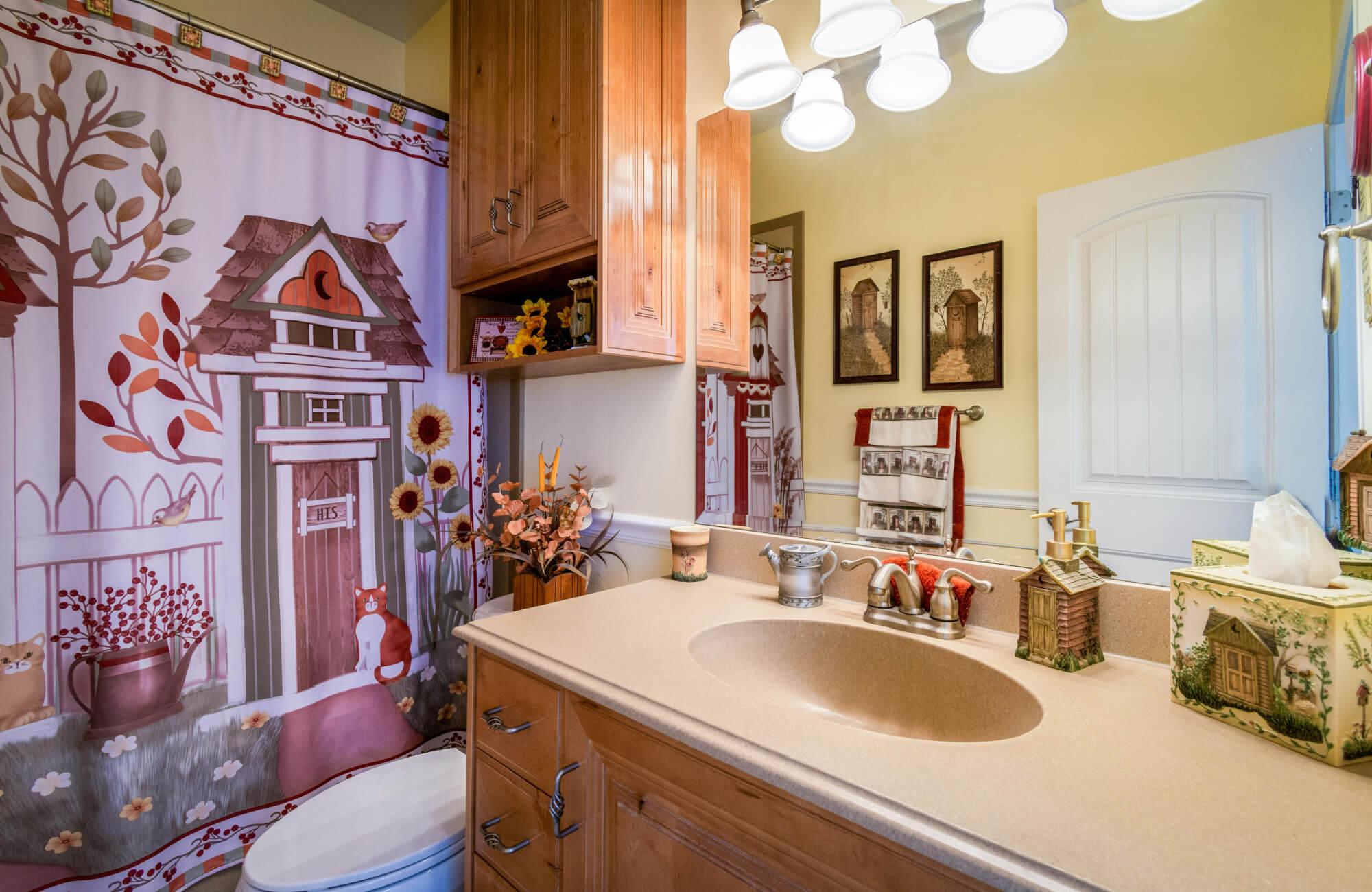 Farmhouse-style bathroom with wood cabinetry, beige countertop, and rustic decor accents, including a patterned shower curtain and floral arrangements.