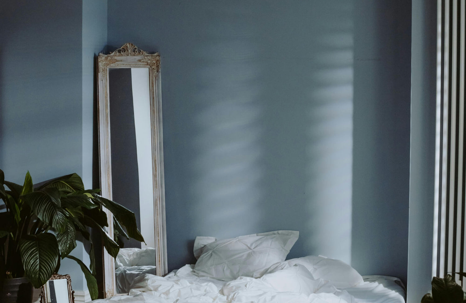 Large floor mirror leaning against a soft blue bedroom wall, reflecting natural light beside an unmade bed and an indoor plant.