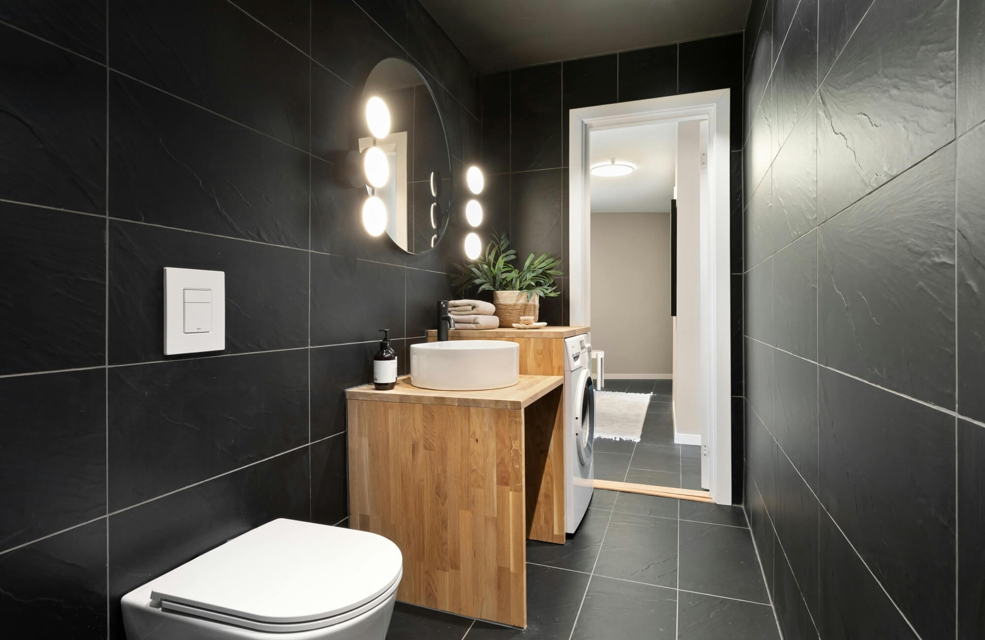 Minimalist bathroom with black wall tiles styled with a wood vanity, round mirror, and soft lighting for balance.