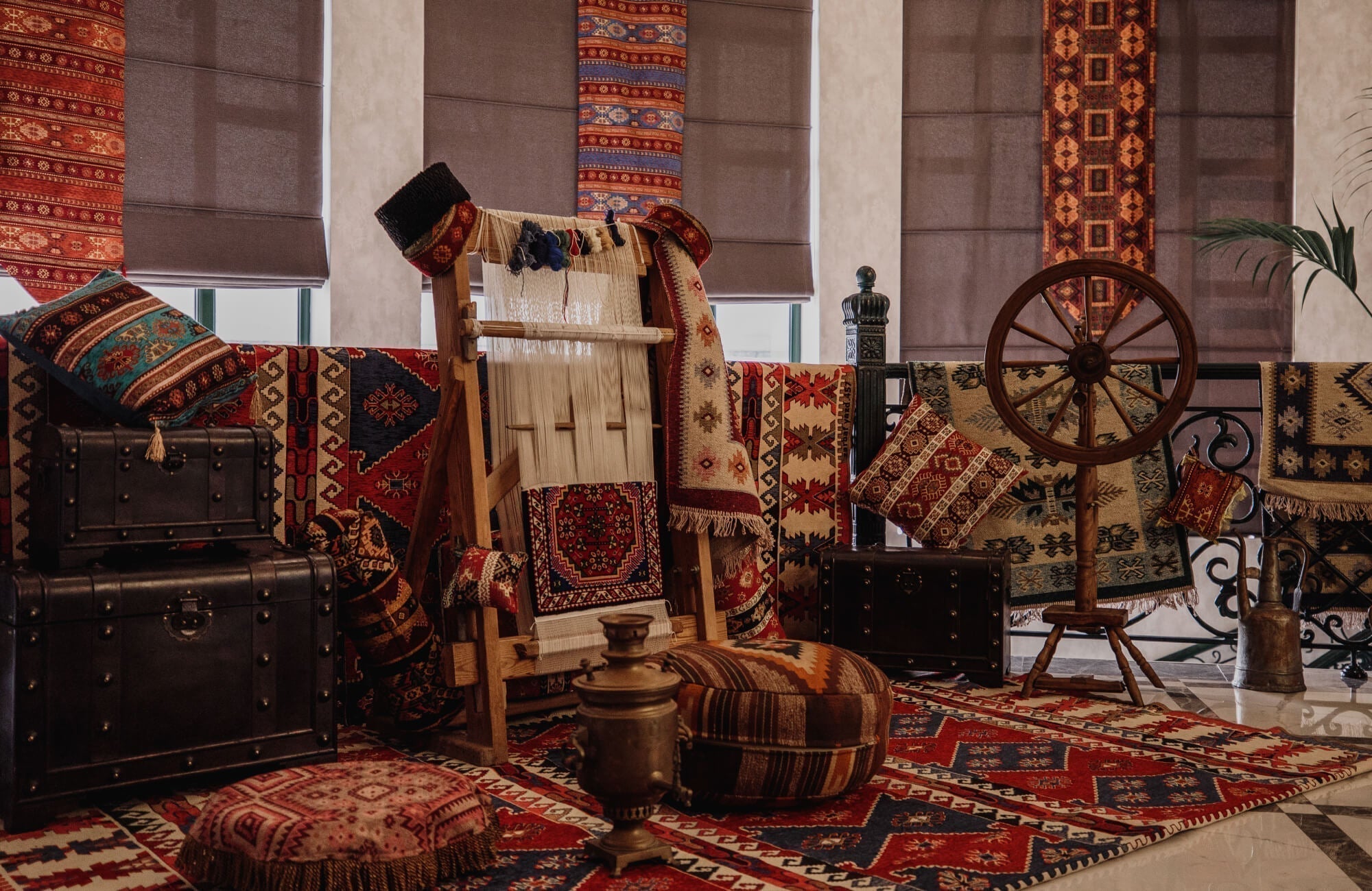 An assortment of richly patterned Persian-style rugs and cushions surrounds a loom and antique decor pieces, showcasing detailed craftsmanship and cultural artistry.