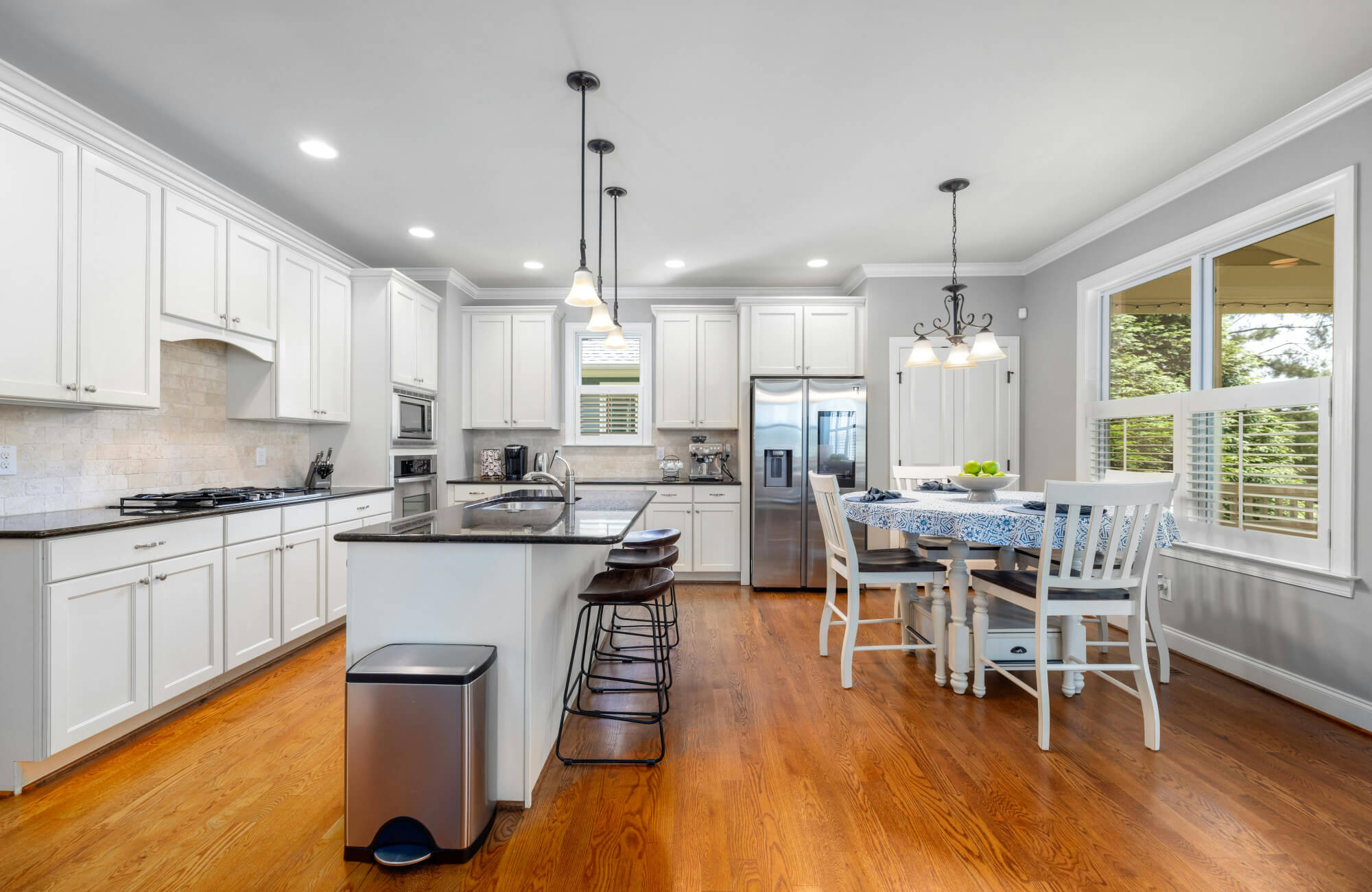 Bright open kitchen with white cabinets, island seating, stainless steel appliances, pendant lighting, and adjacent dining nook with table.