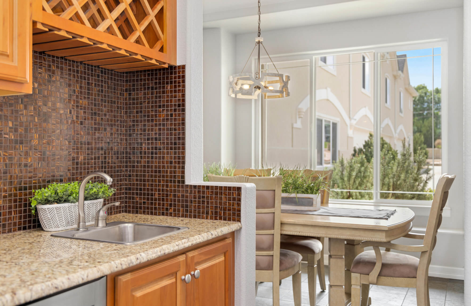 Brown mosaic tile backsplash with small square patterns behind a stainless sink and granite countertop in a bright kitchen and dining area.