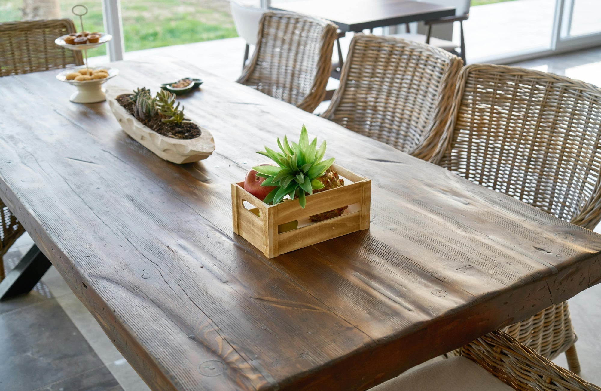 Rectangular wooden dining table with textured surface paired with woven chairs in a bright indoor-outdoor setting.