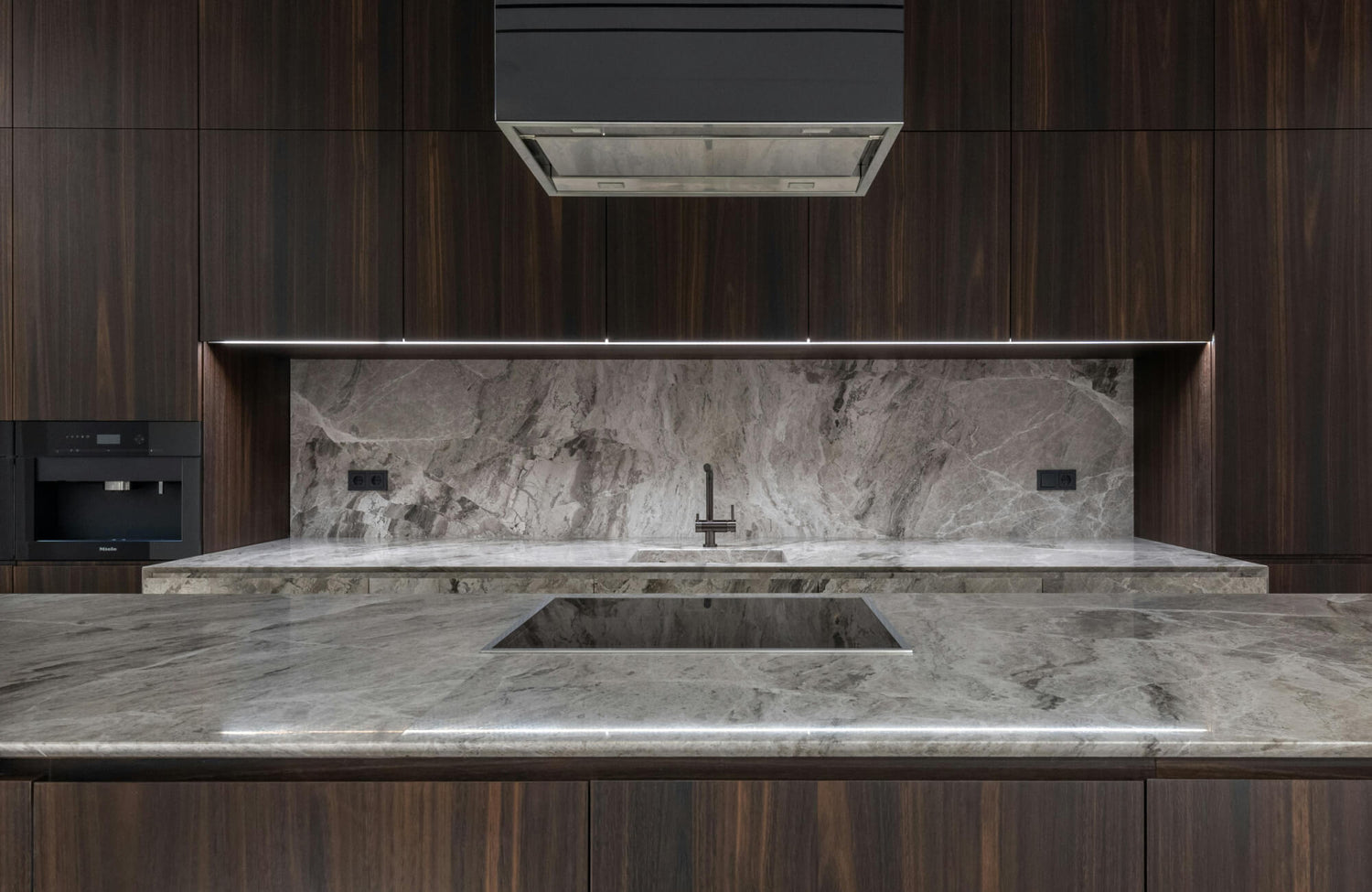 Full-height stone backsplash with dramatic gray veining adds depth and continuity behind a modern cooktop and sink area.