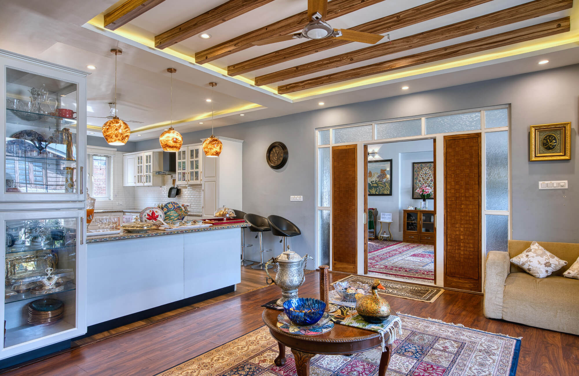 Modern open living space with layered rugs, warm wood ceiling beams, pendant lighting, and a connected kitchen and seating area.