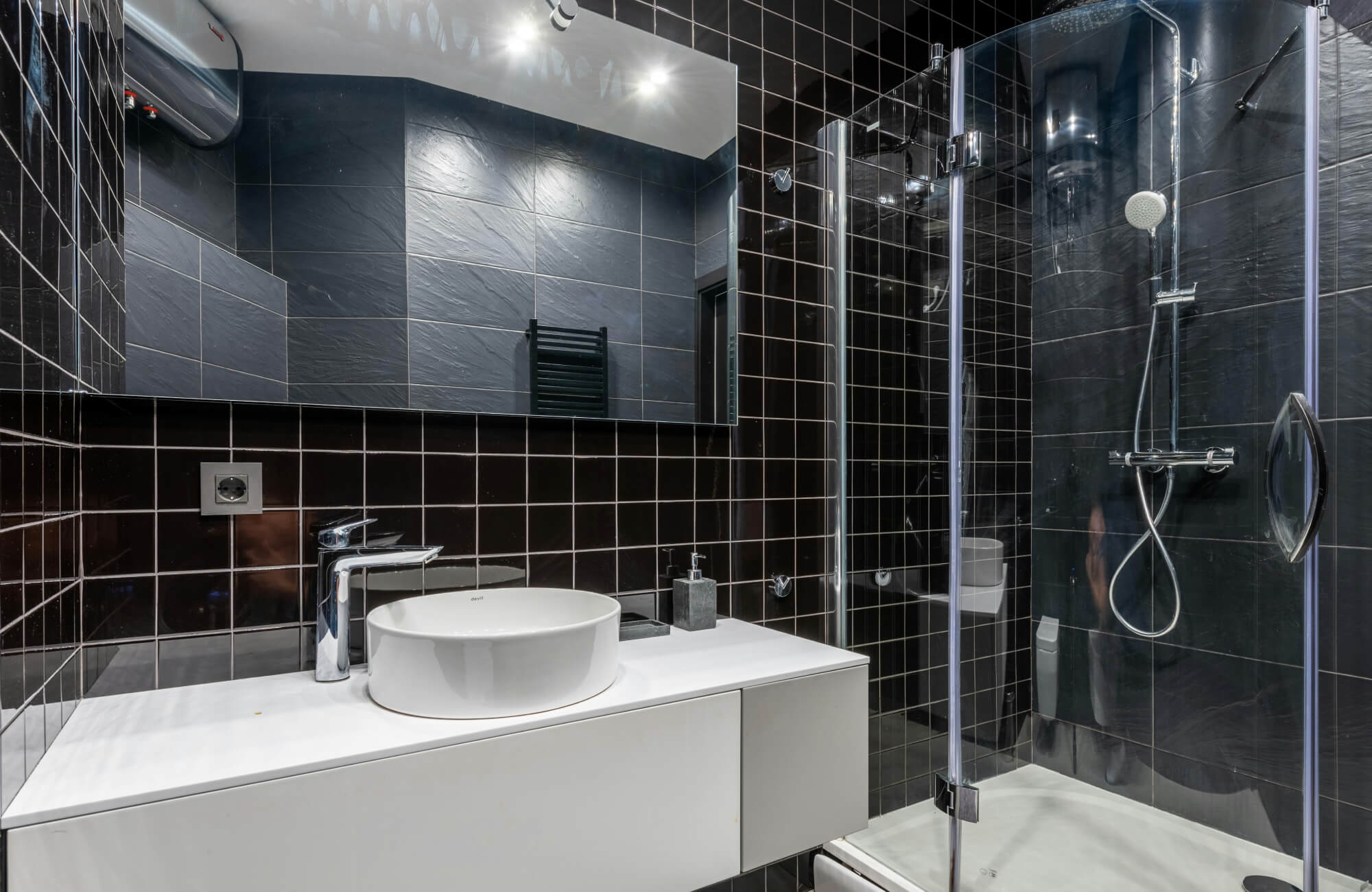 Modern bathroom with black tiled shower and white vanity featuring reflective surfaces and bright overhead lighting.