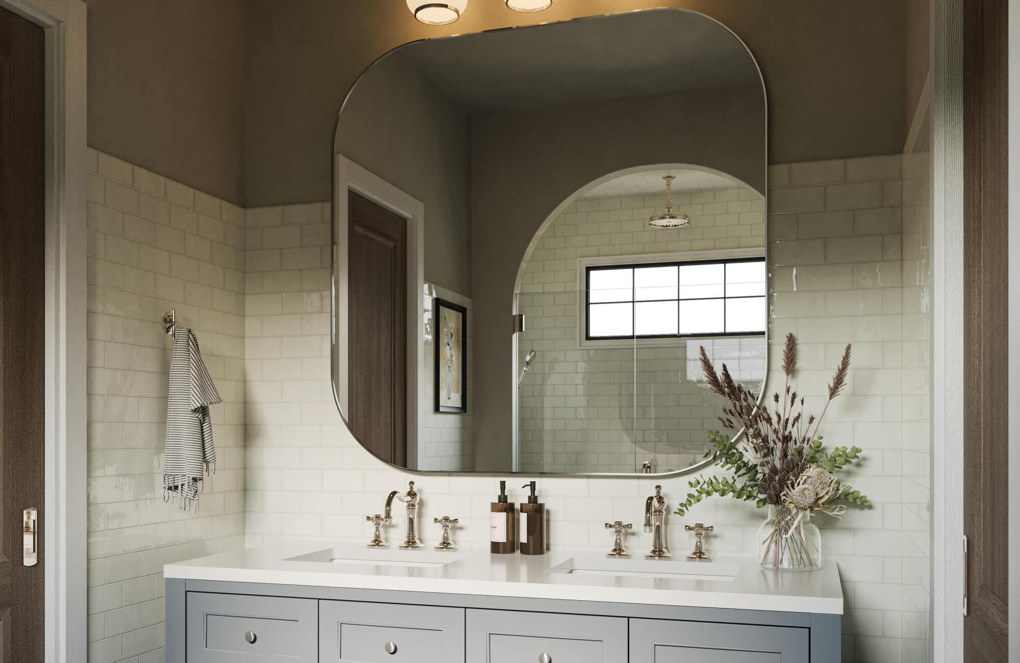 A bright, elegant bathroom features a soft gray double vanity topped with polished fixtures, glossy subway-tiled walls, and a striking large wall mirror that reflects the arched shower entry and enhances the room’s airy, sophisticated ambiance.