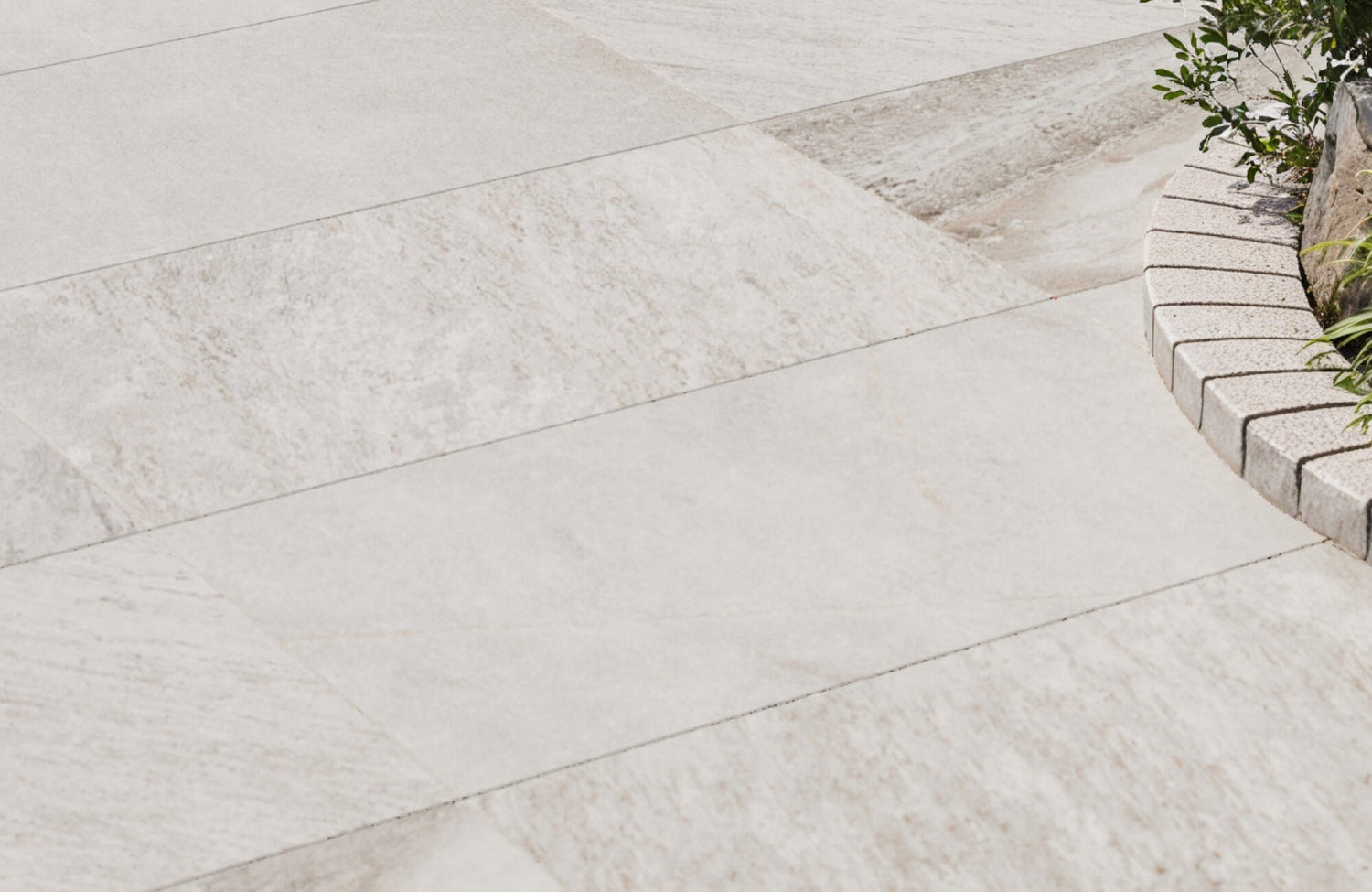 Elegant light beige porcelain outdoor paver tiles with subtle stone-like texture, arranged beside a curved garden border with greenery.