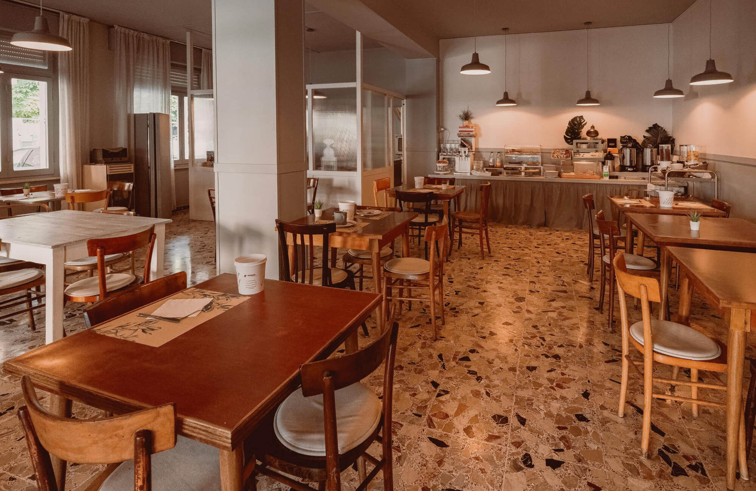 Cozy café with mismatched wooden chairs, terrazzo floors, pendant lighting, and a self-serve breakfast bar along the back wall.