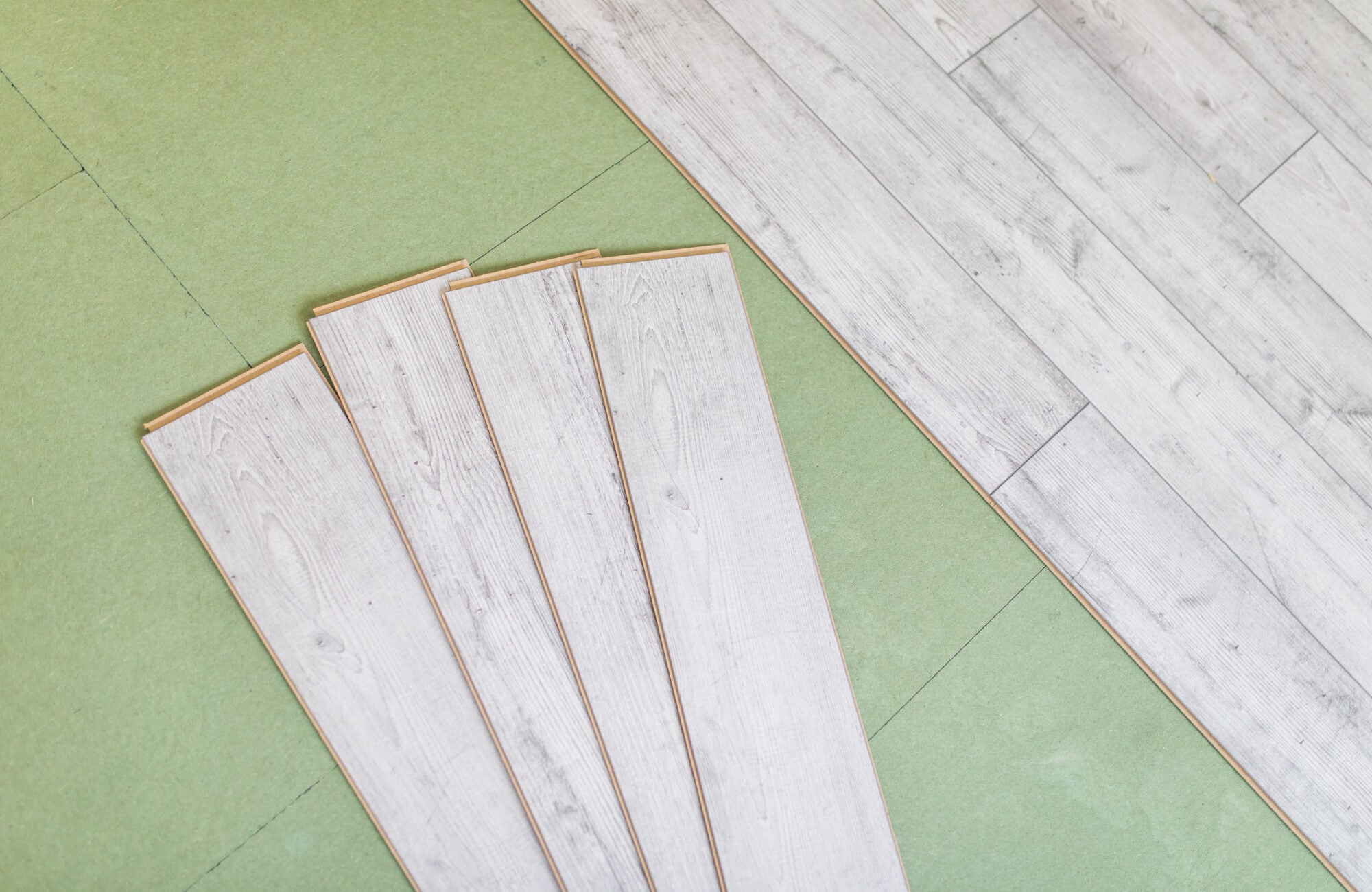 Light gray vinyl plank flooring is being installed over a green underlayment, with several planks laid out in preparation.