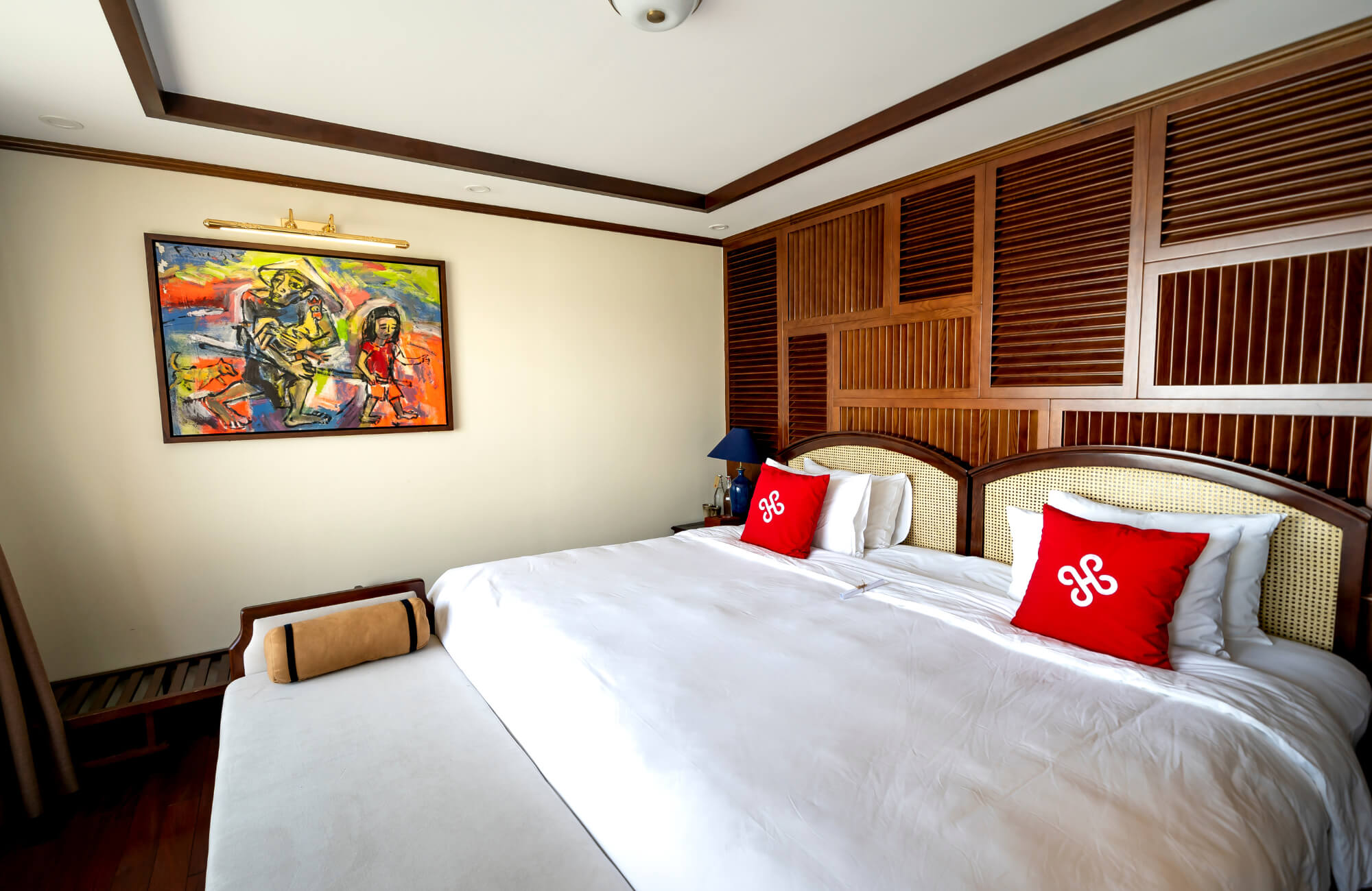 A modern bedroom with a colorful framed painting illuminated by a brass picture light above a large bed with red accent pillows.