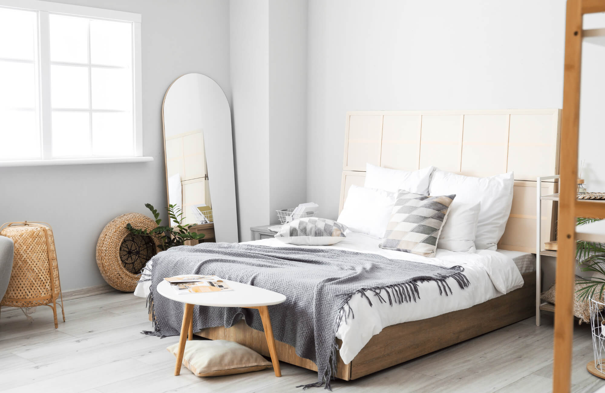 Modern bedroom with a full-length arched mirror, neutral bedding, light wood furniture, and natural woven accents in soft daylight.