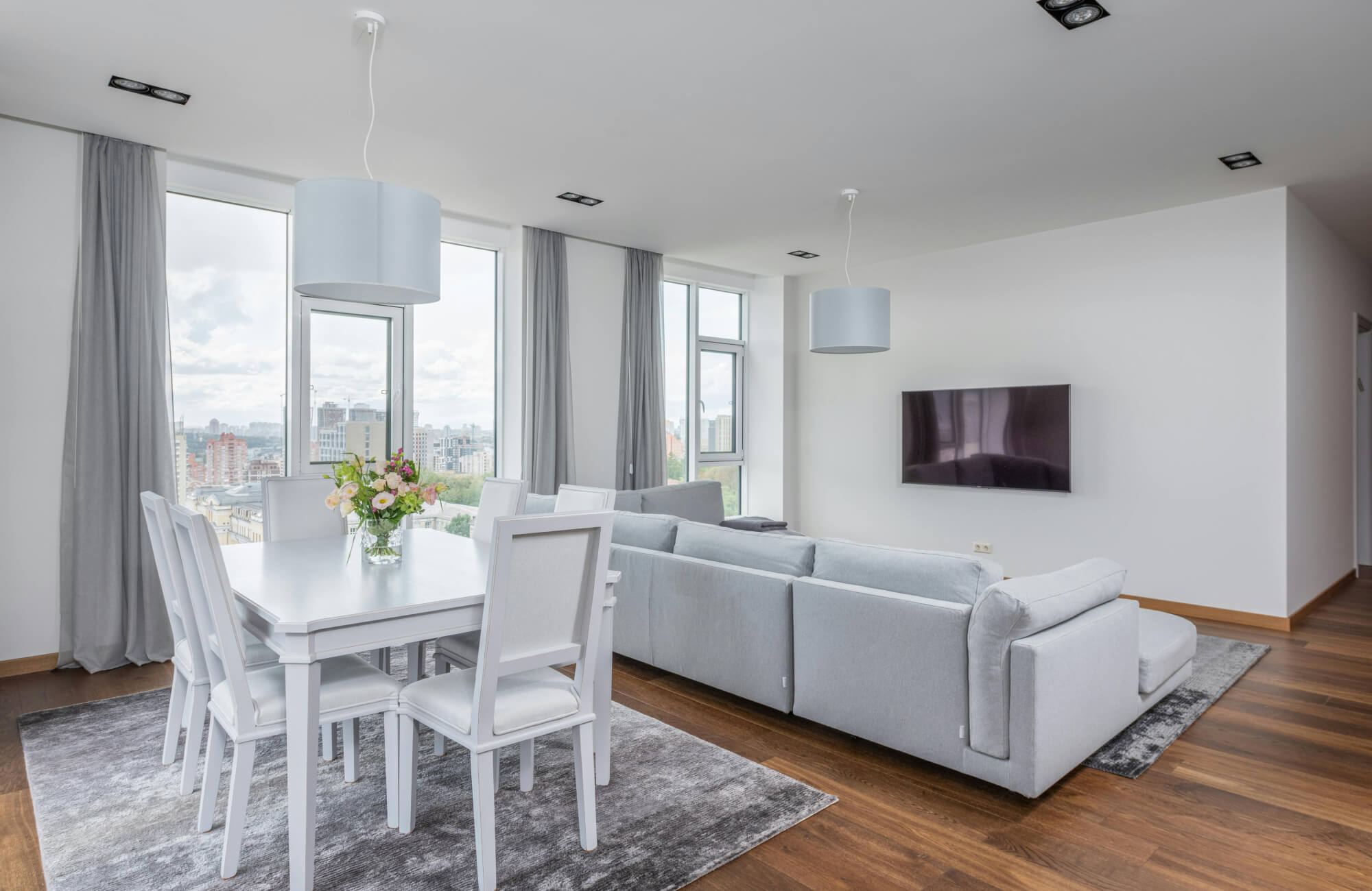 Bright dining room with a soft gray rug beneath a white table, paired with large windows and minimal decor.