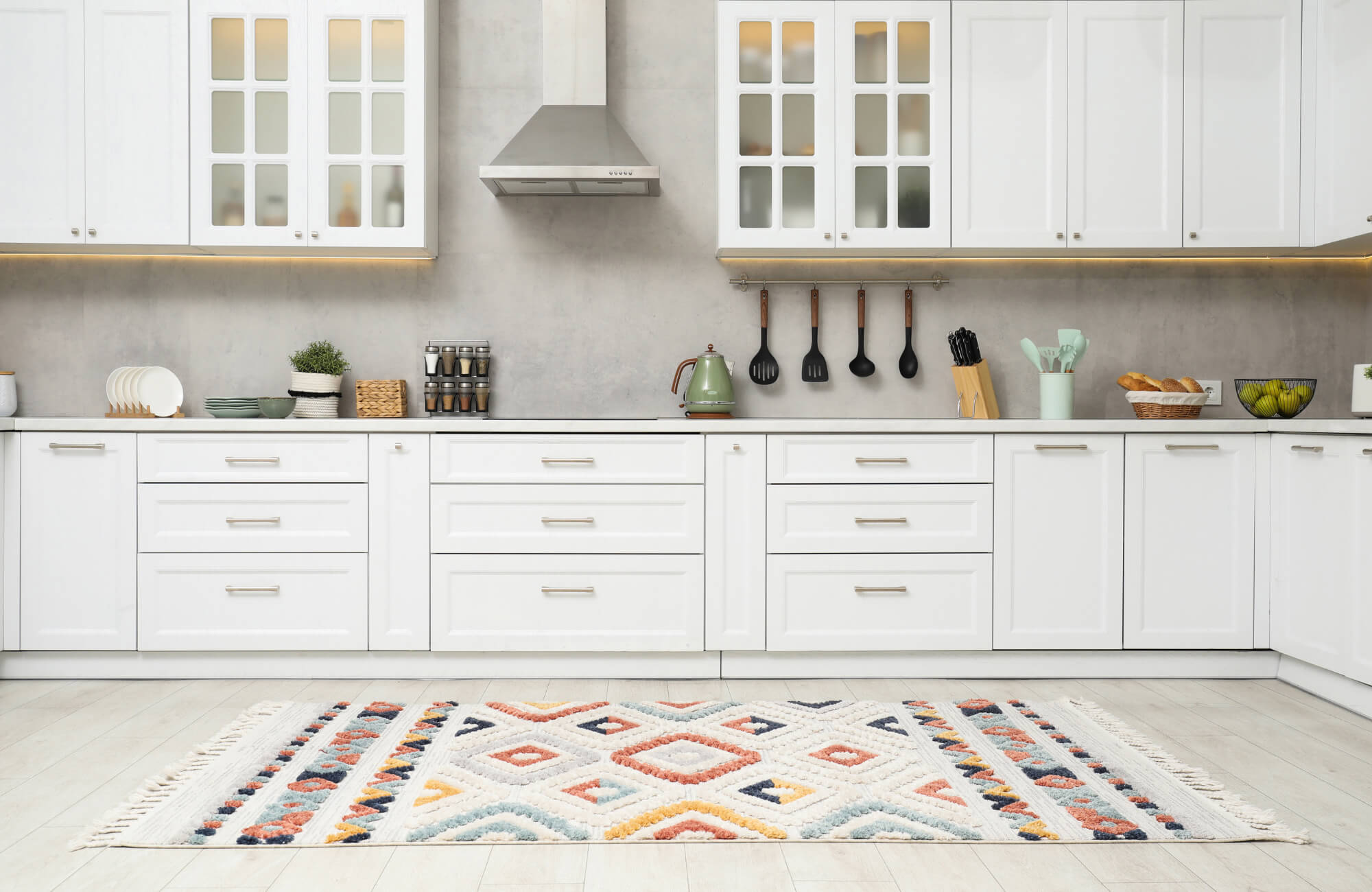 Modern white kitchen with cabinets, stainless steel hood, and a colorful geometric rug on light wood flooring.