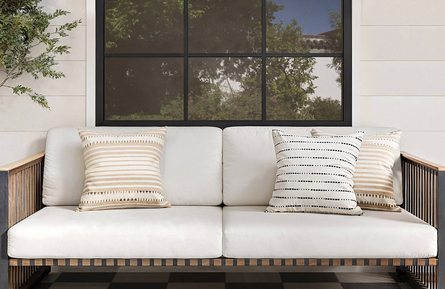 Outdoor sofa with white cushions styled with three square throw pillows in beige, cream, and black striped patterns beneath a large black-framed window.