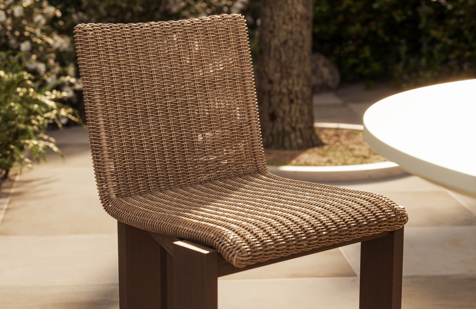 Close-up of a woven brown outdoor dining chair with a curved seat and dark wood legs on a softly sunlit patio beside a round table.